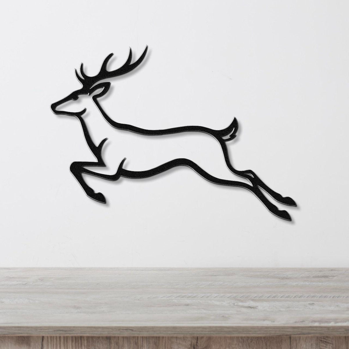 Black metal wall art of a leaping deer silhouette with antlers, mounted on a white wall above a light wood surface.