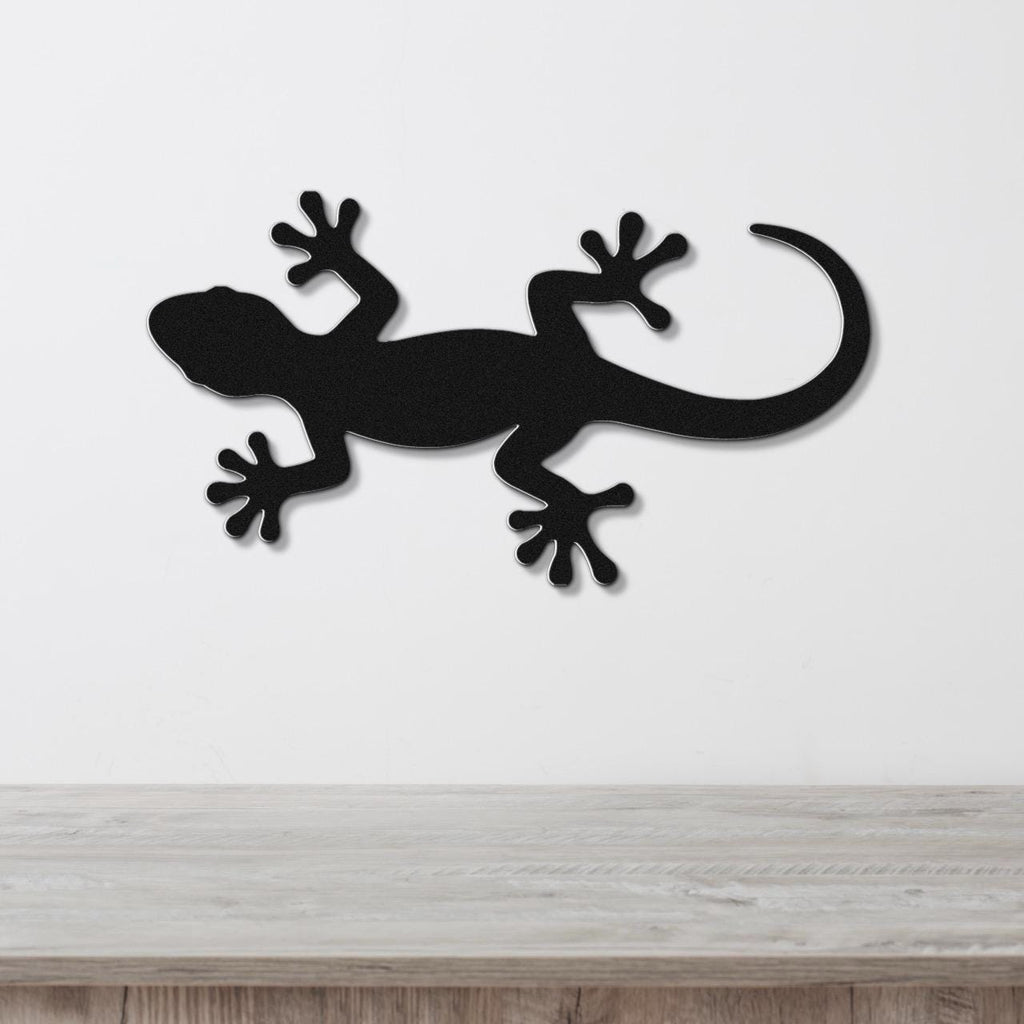 Black metal gecko wall art silhouette with detailed feet and curved tail, mounted on a white wall above a light wood surface.