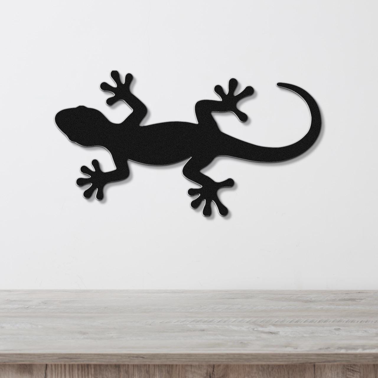 Black metal gecko wall art silhouette with detailed feet and curved tail, mounted on a white wall above a light wood surface.