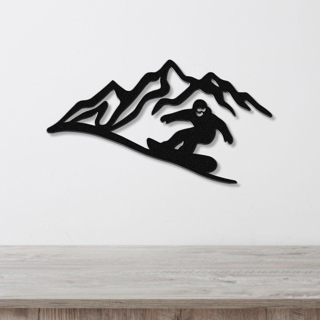 Black metal wall art featuring a snowboarder silhouette descending a mountain range, mounted on a white wall above a light wood surface.