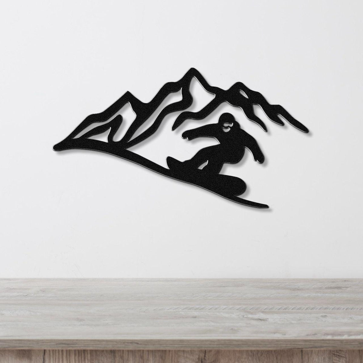 Black metal wall art featuring a snowboarder silhouette descending a mountain range, mounted on a white wall above a light wood surface.