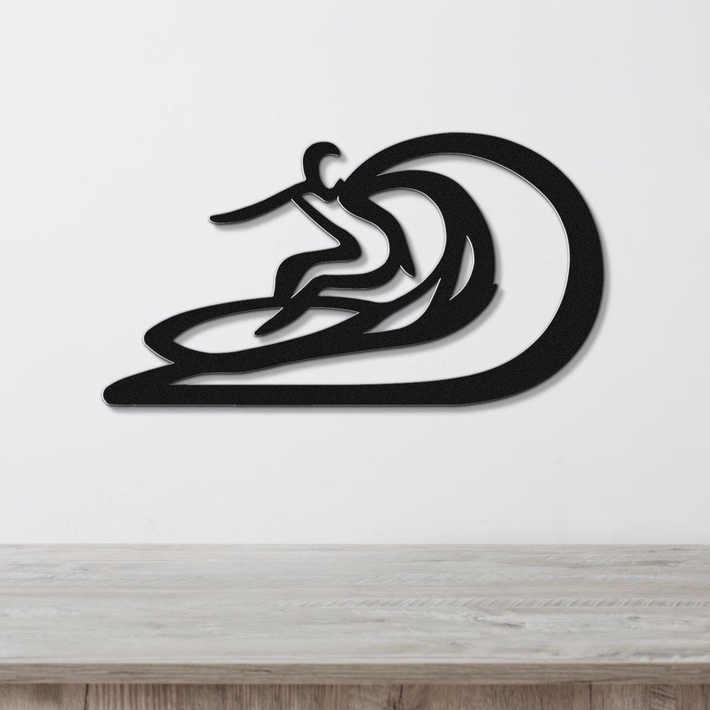 Black metal wall sculpture of a surfer riding a wave, with layered cutout details, mounted on a white wall above a wooden surface.