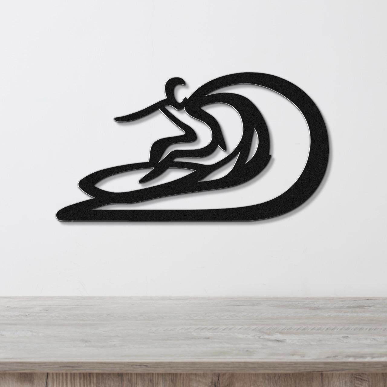 Black metal wall sculpture of a surfer riding a wave, with layered cutout details, mounted on a white wall above a wooden surface.