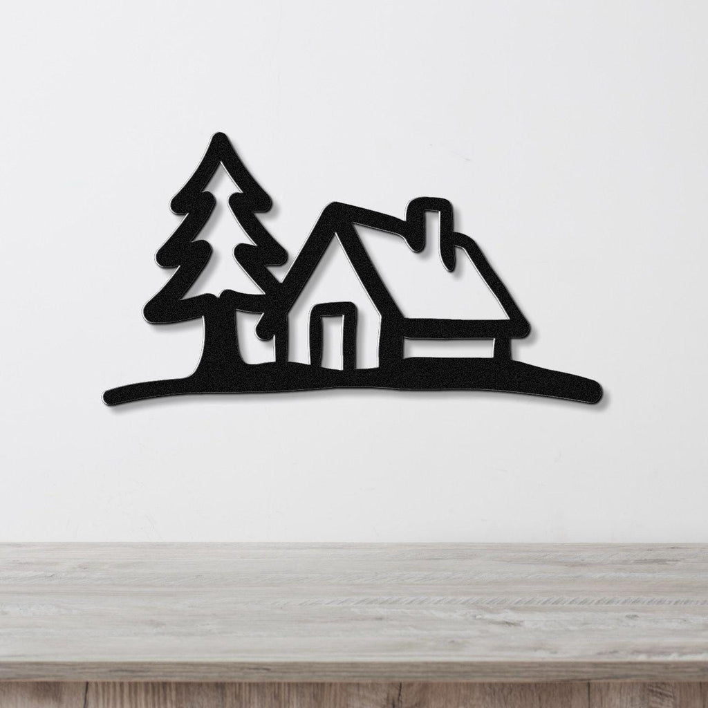 Black metal wall art cutout of a cabin and pine tree silhouette, mounted on a white wall above a wooden surface.