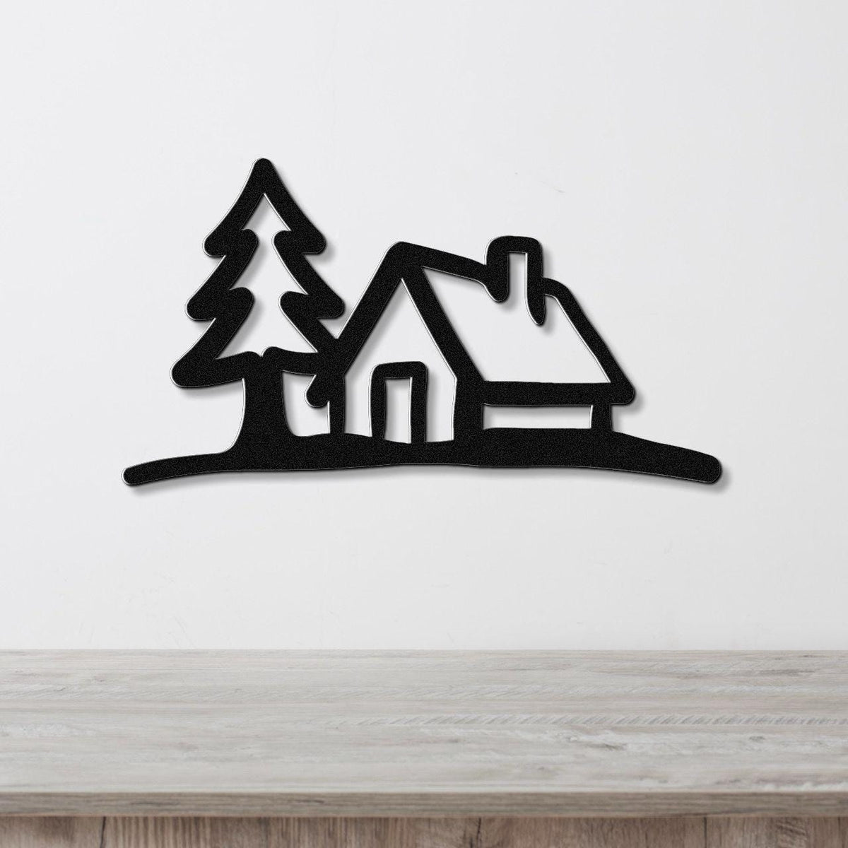 Black metal wall art cutout of a cabin and pine tree silhouette, mounted on a white wall above a wooden surface.