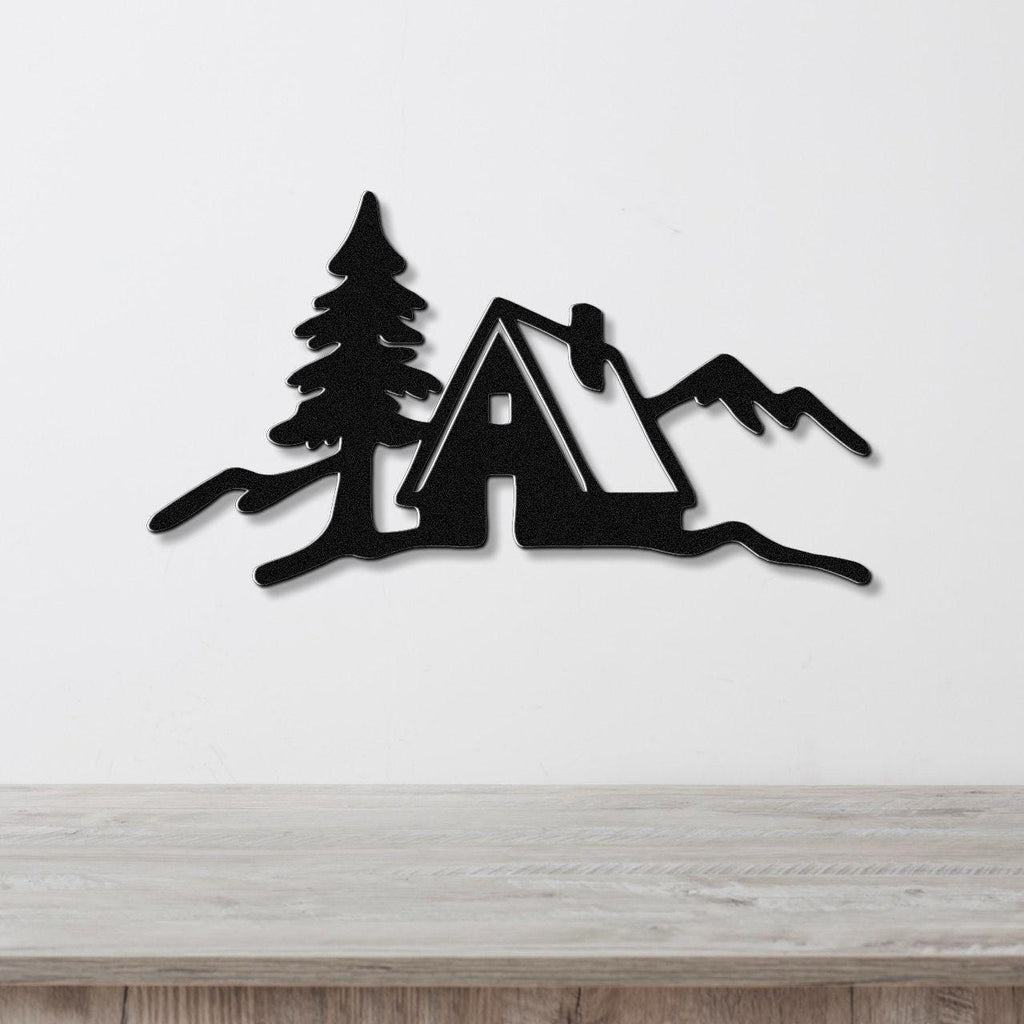 Black metal wall art silhouette of a cabin, pine tree, and mountains, mounted on a white wall above a light wood counter.