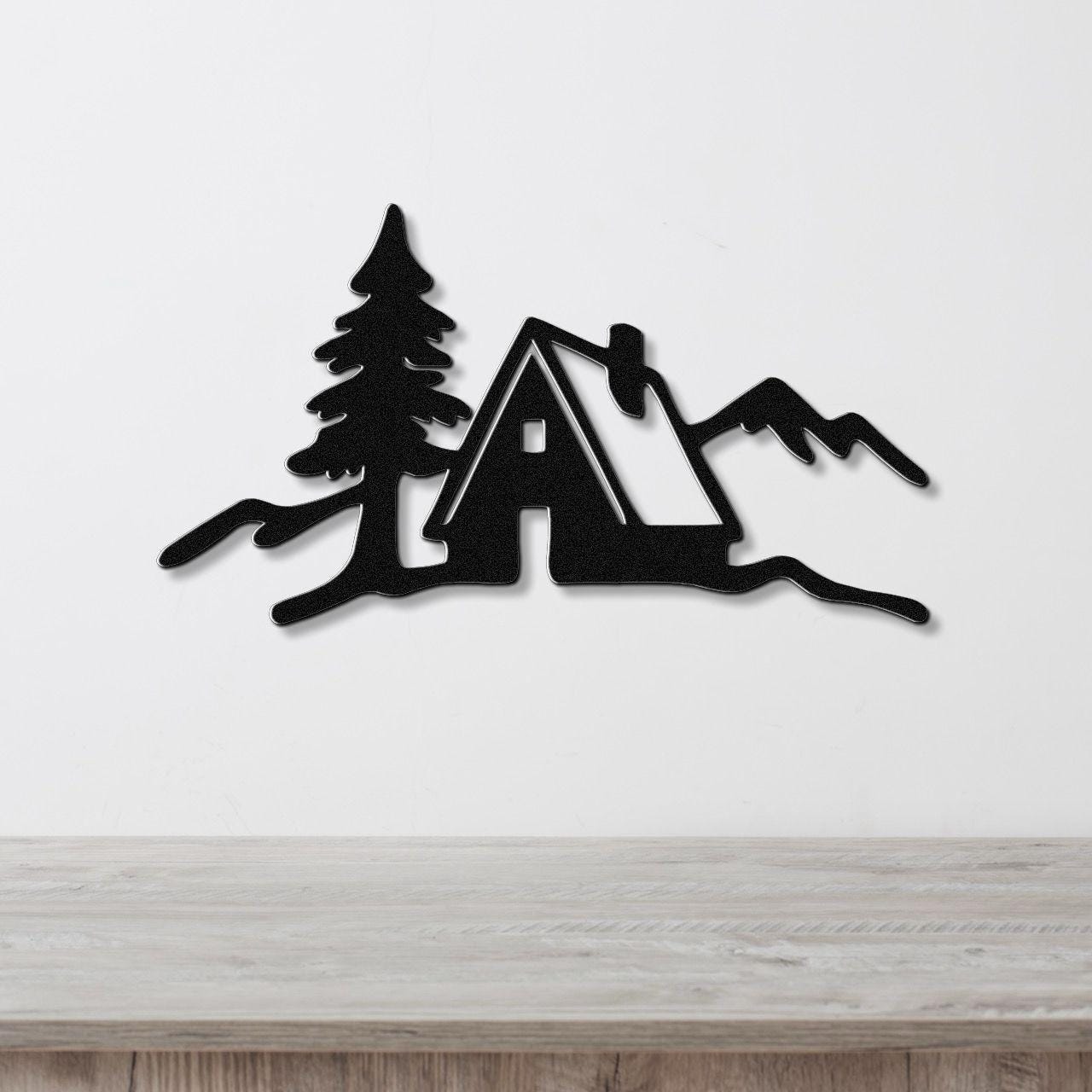 Black metal wall art silhouette of a cabin, pine tree, and mountains, mounted on a white wall above a light wood counter.
