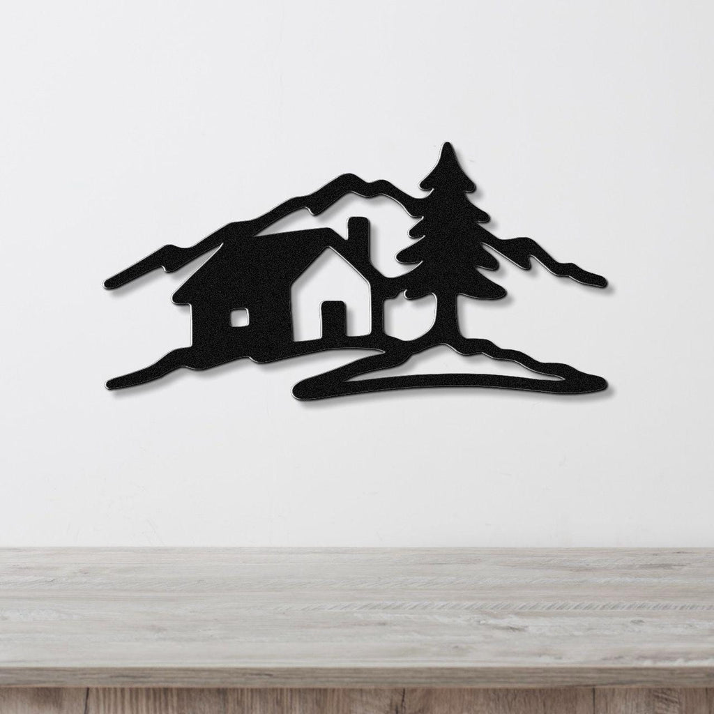 Black metal wall art cutout of a cabin, pine tree, and mountains, mounted on a white wall above a wooden surface.