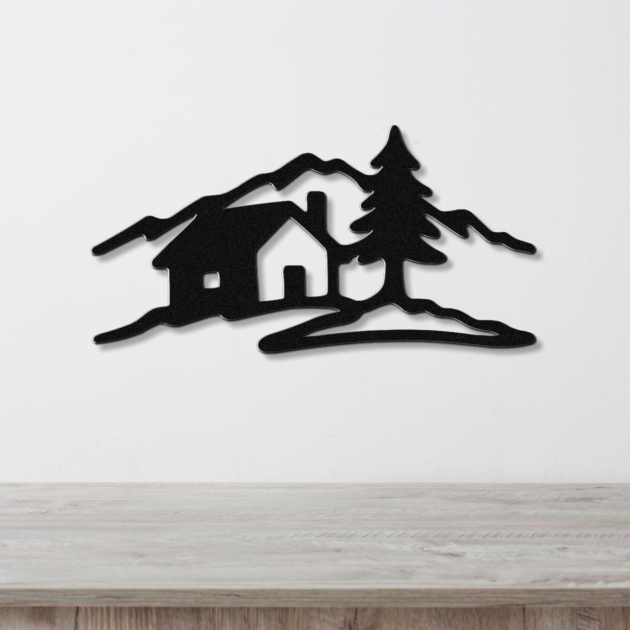 Black metal wall art cutout of a cabin, pine tree, and mountains, mounted on a white wall above a wooden surface.