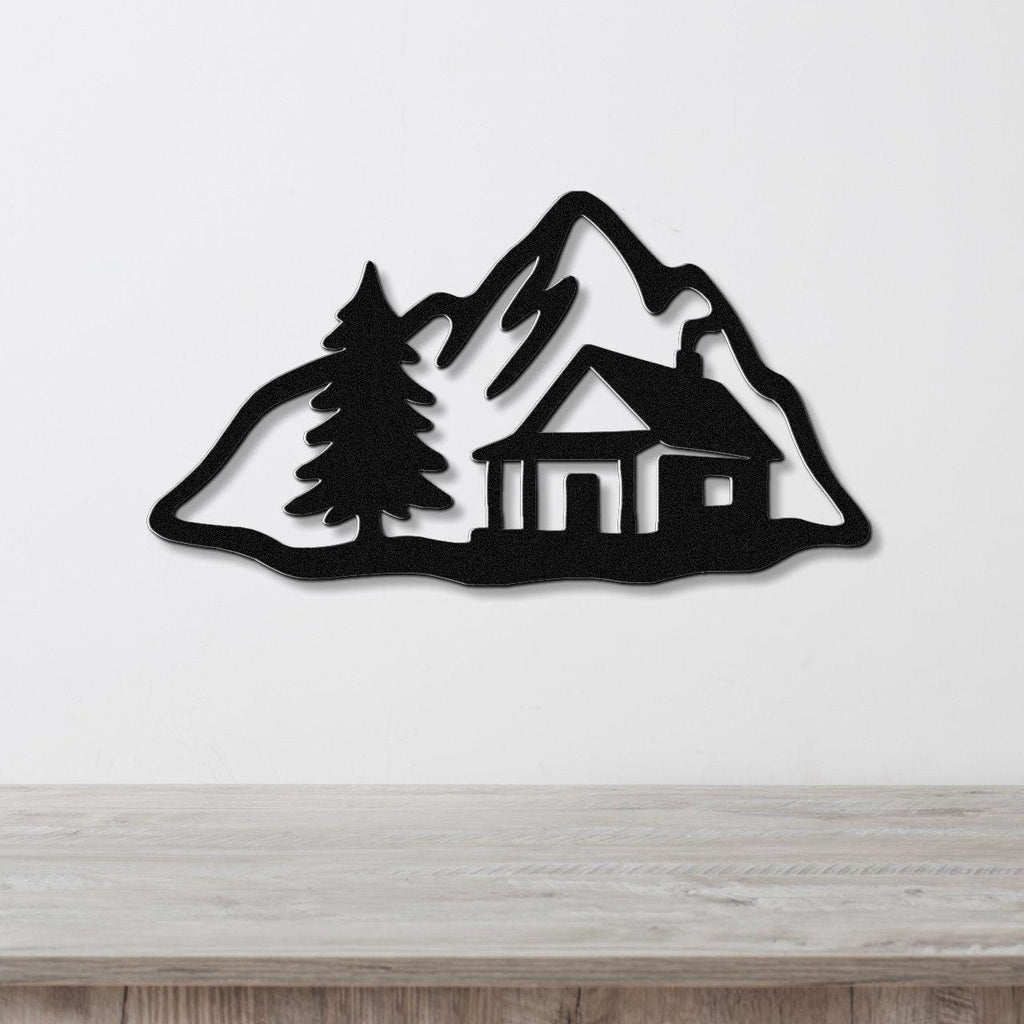 Black metal wall art silhouette of a mountain cabin with a pine tree, mounted on a white wall above a wooden surface.