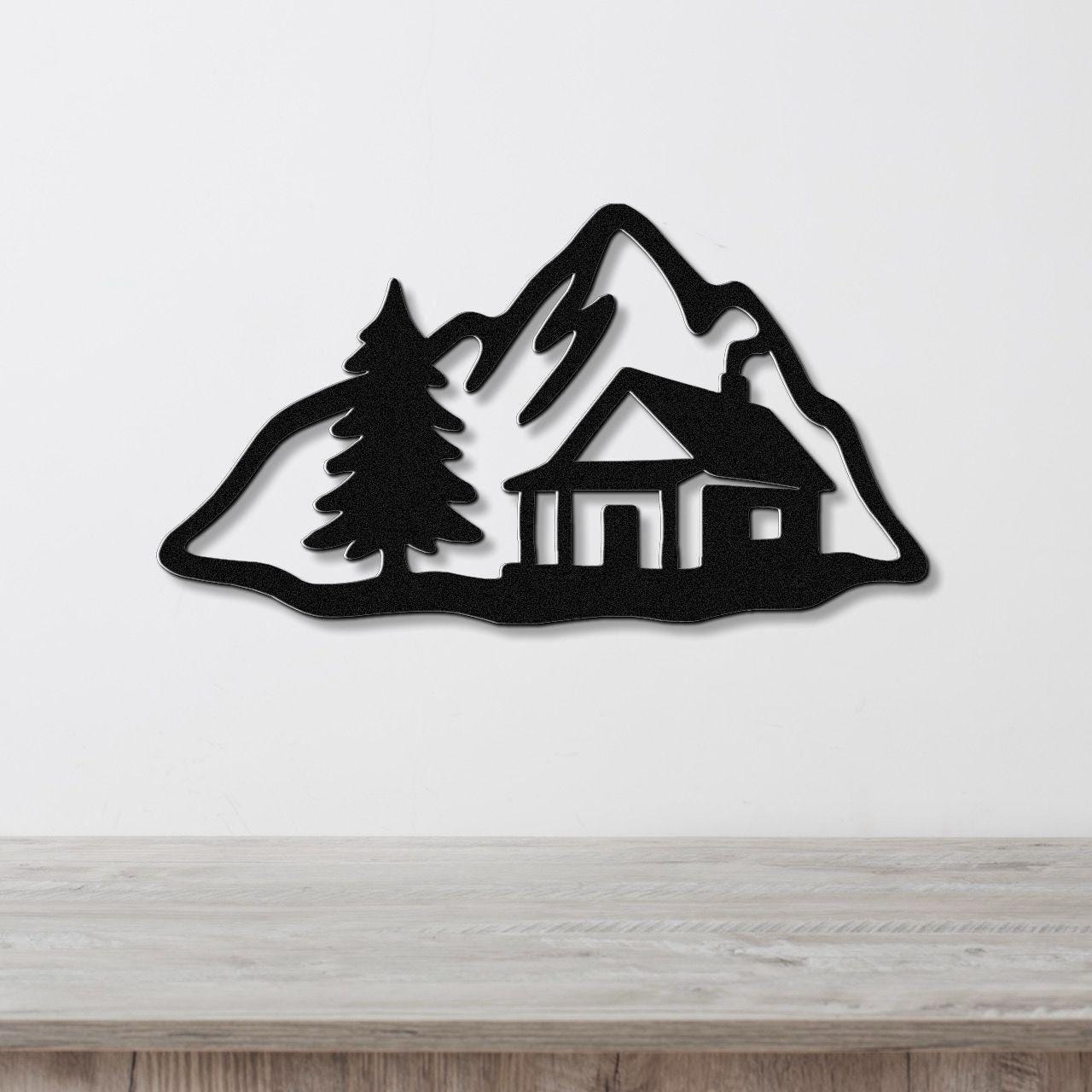 Black metal wall art silhouette of a mountain cabin with a pine tree, mounted on a white wall above a wooden surface.