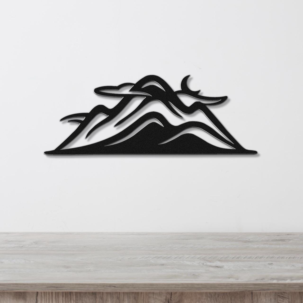 Black metal wall art featuring a layered mountain silhouette with internal cutout details, mounted on a white wall above a light wood surface.