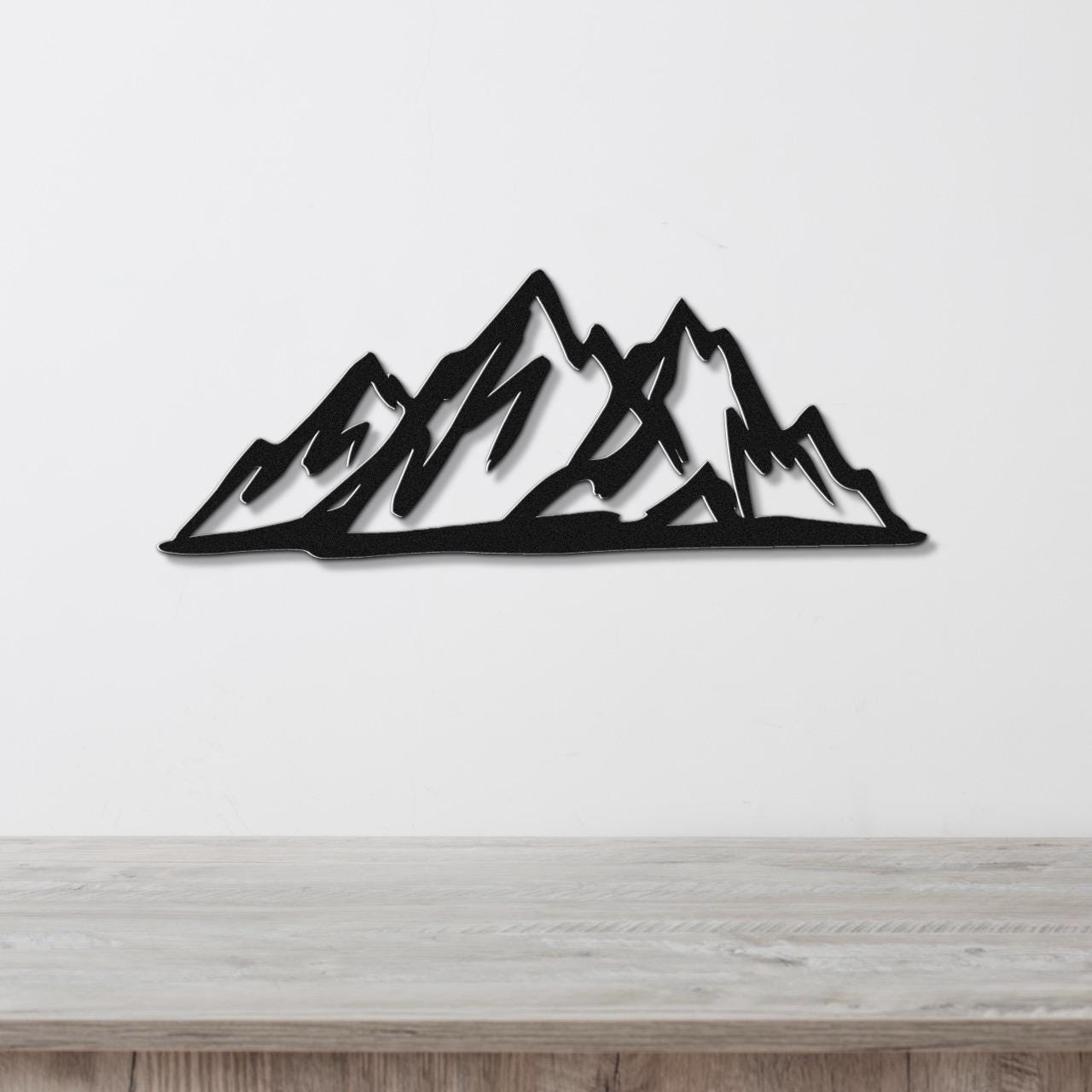 Black metal mountain silhouette wall art with layered peaks, mounted on a white wall above a light wood surface.