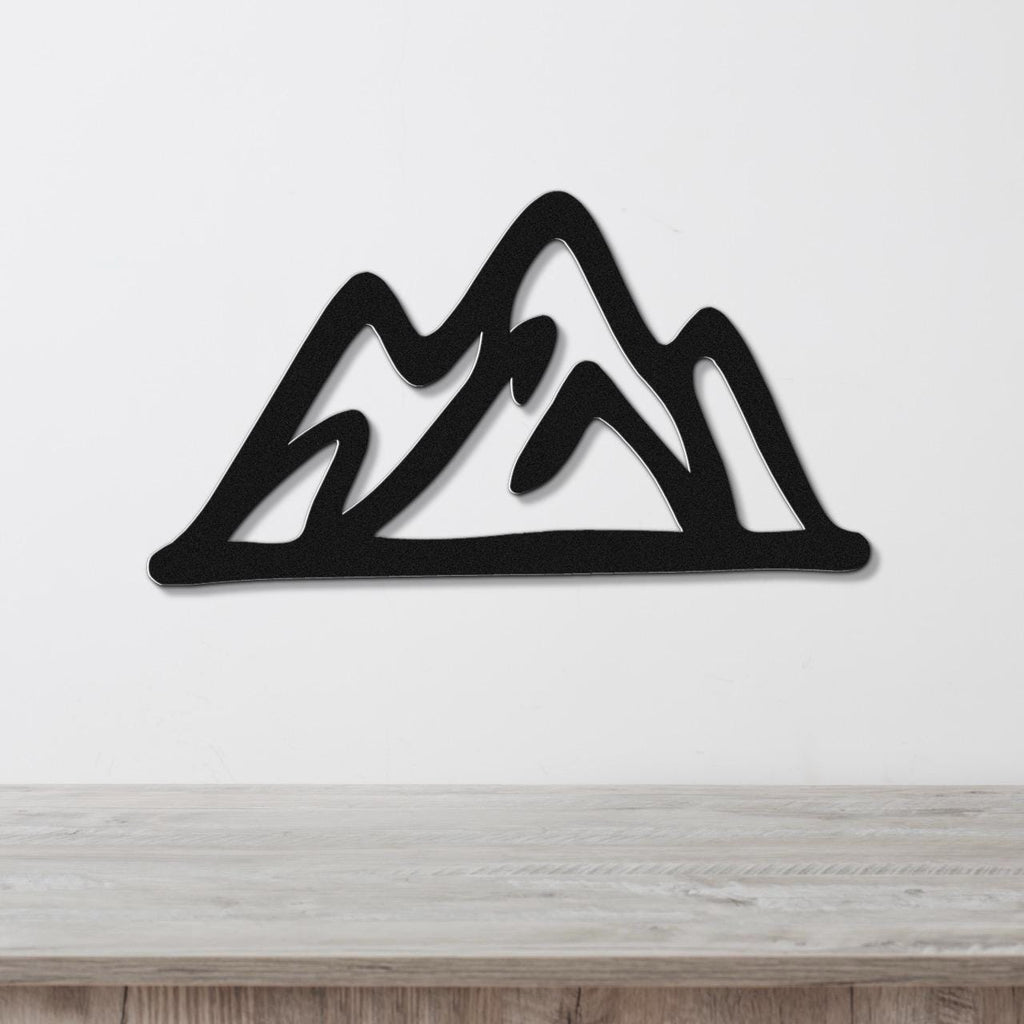 Black metal mountain wall art with layered peak cutouts, mounted on a white wall above a light wood surface.