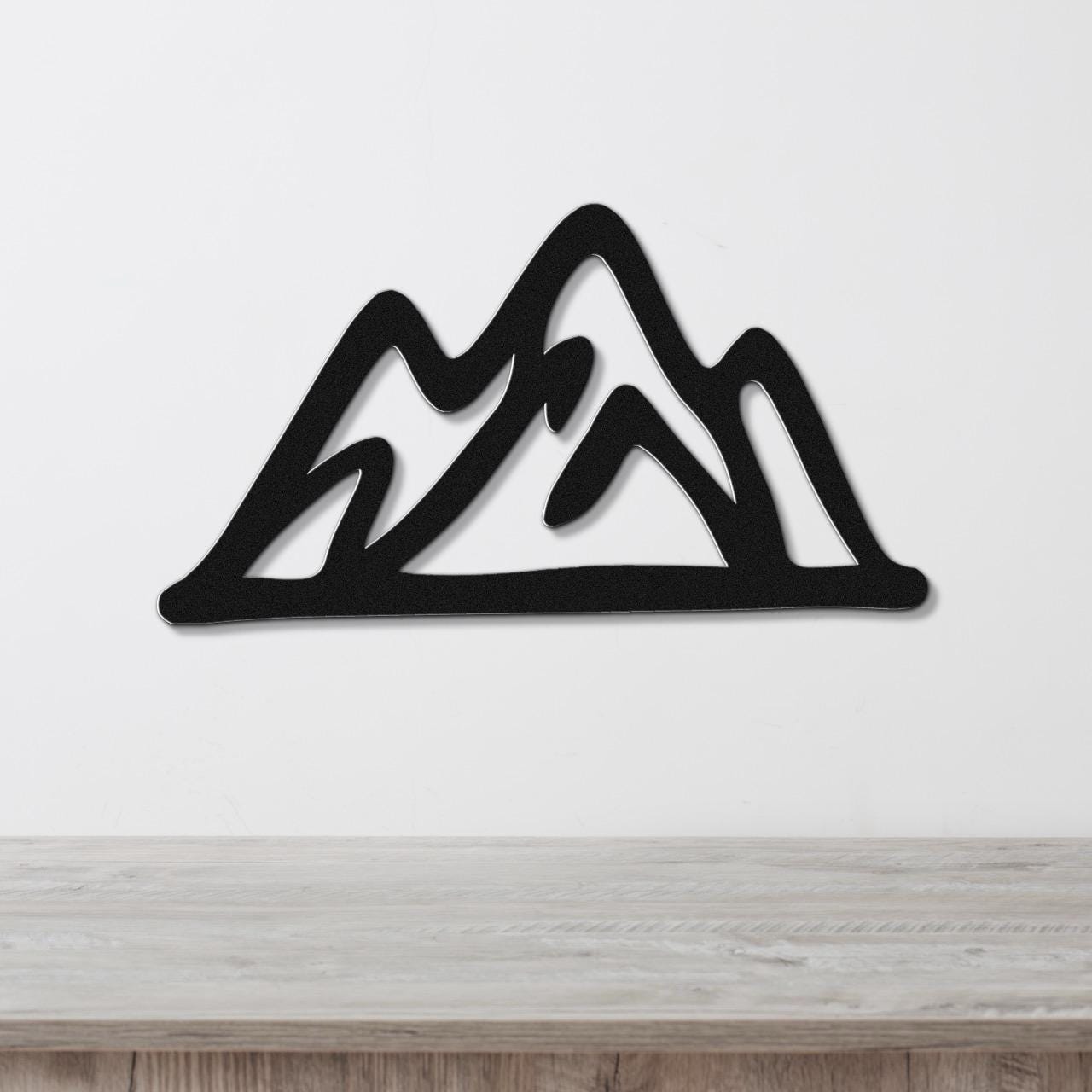 Black metal mountain wall art with layered peak cutouts, mounted on a white wall above a light wood surface.