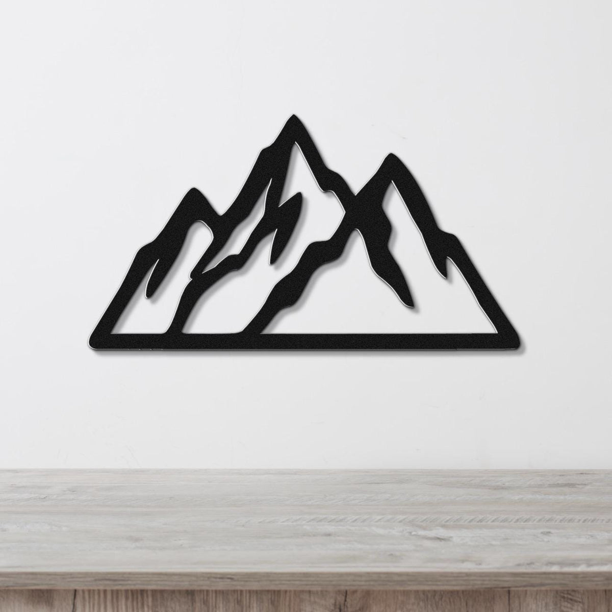 Black metal mountain wall art with layered peaks and internal cutouts, mounted on a white wall above a light wood surface.