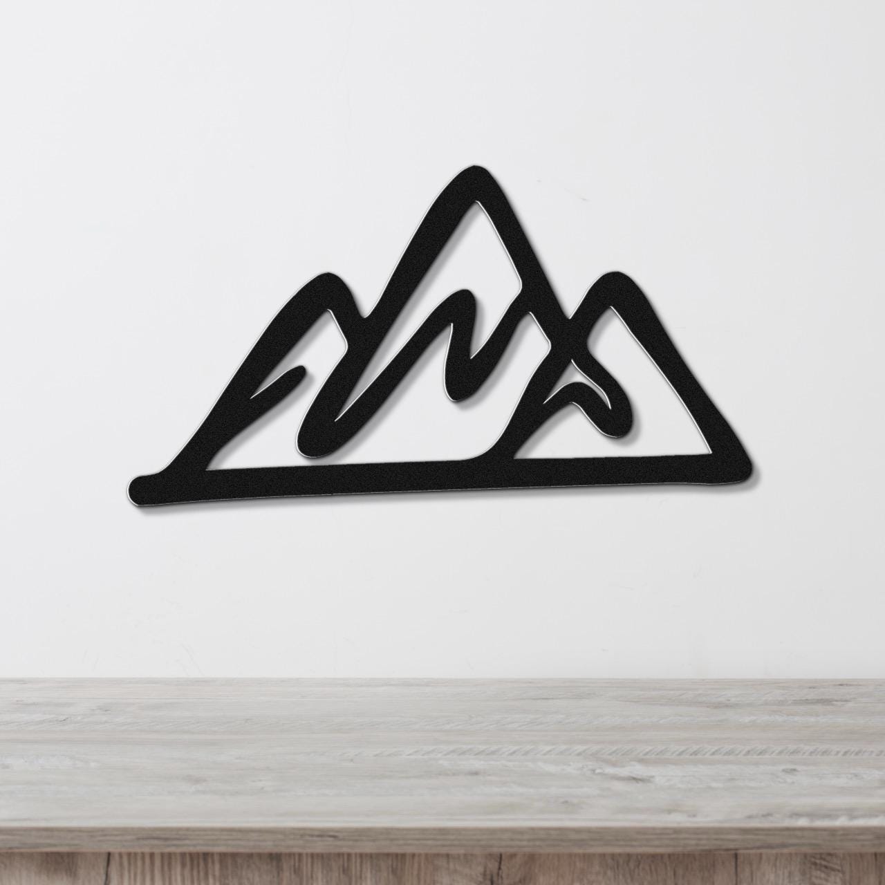 Black metal mountain silhouette wall art with layered peaks, mounted on a white wall above a light wood surface.