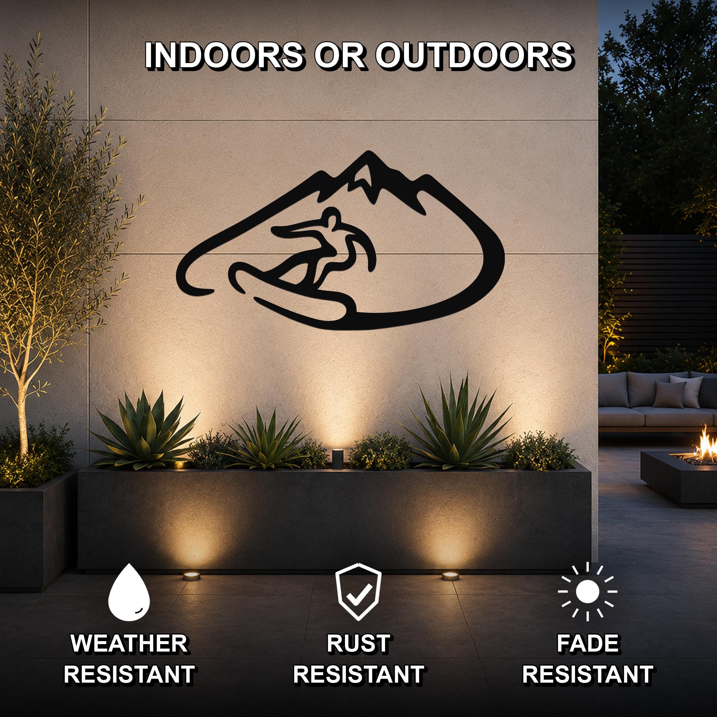 Black metal wall art featuring a snowboarder silhouette inside a mountain outline, mounted on an outdoor wall above a planter in a modern patio setting.