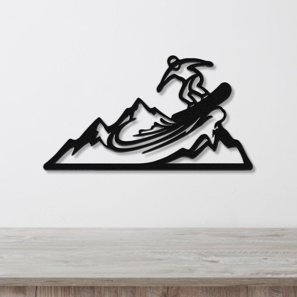 Black metal wall art featuring a snowboarder silhouette mid-air over mountain peaks, mounted on a white wall above a wooden surface.