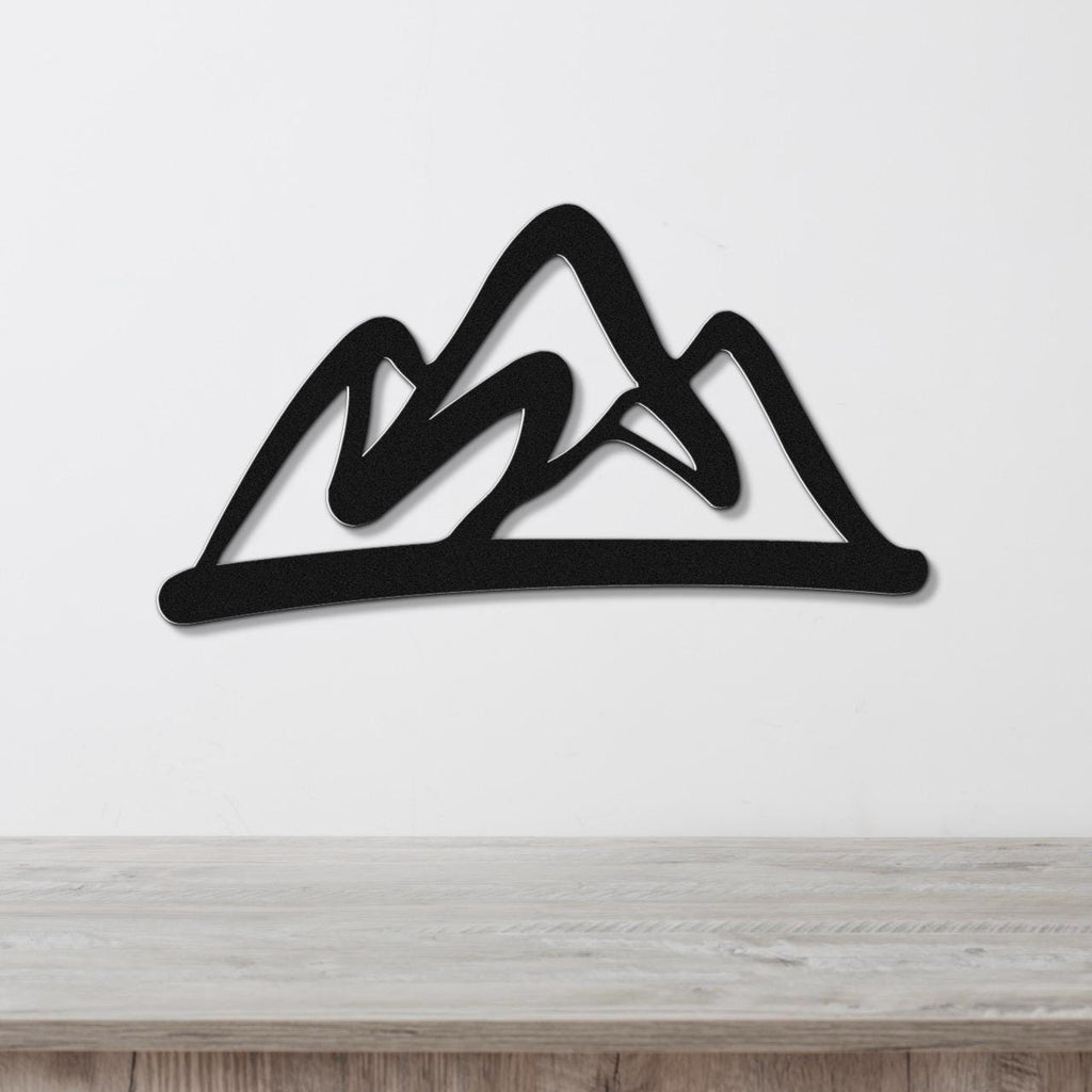 Black metal wall art with a bold mountain silhouette design, mounted on a white wall above a light wood surface.