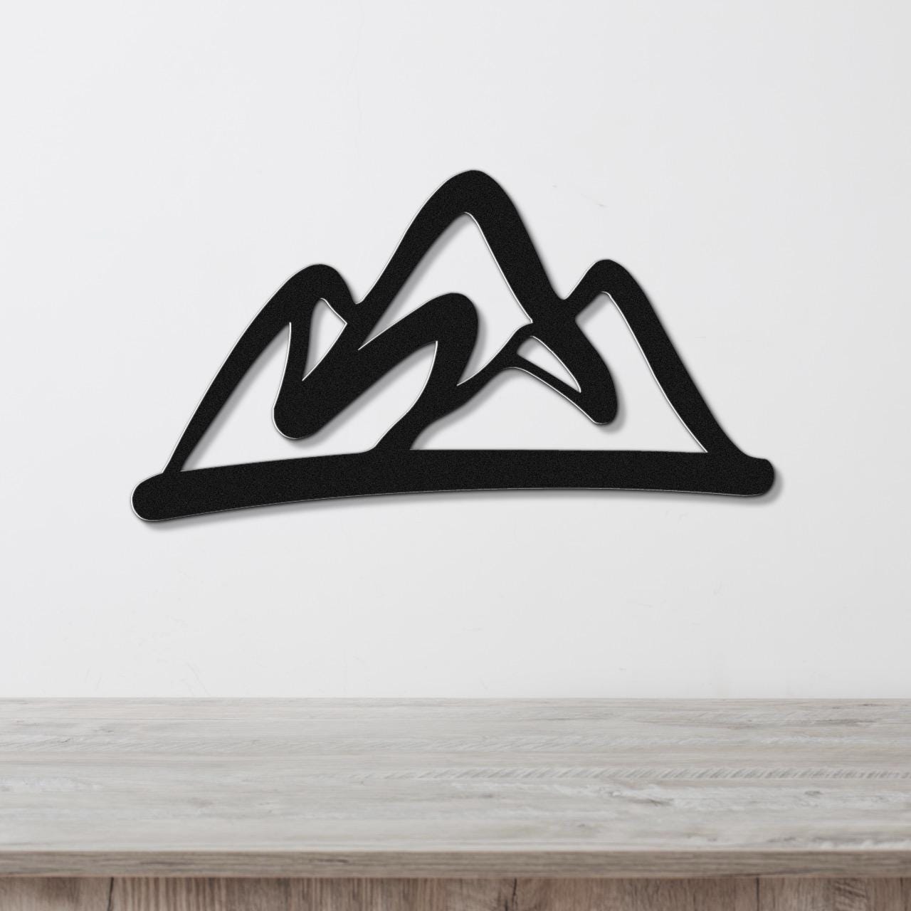 Black metal wall art with a bold mountain silhouette design, mounted on a white wall above a light wood surface.