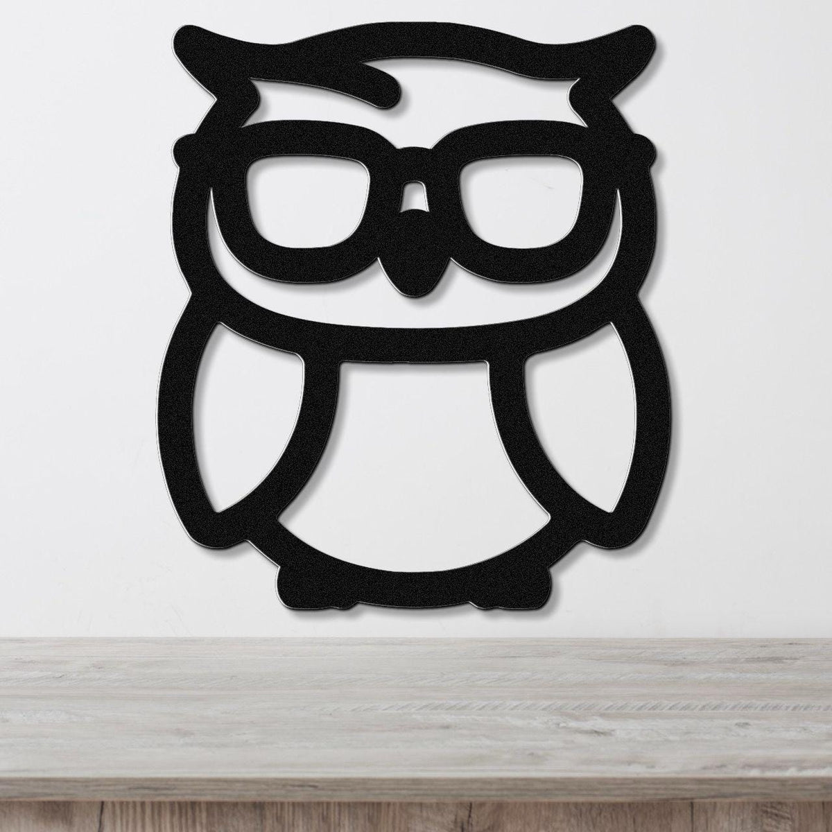 Black metal owl wall art with glasses silhouette, mounted on a white wall above a wooden surface.