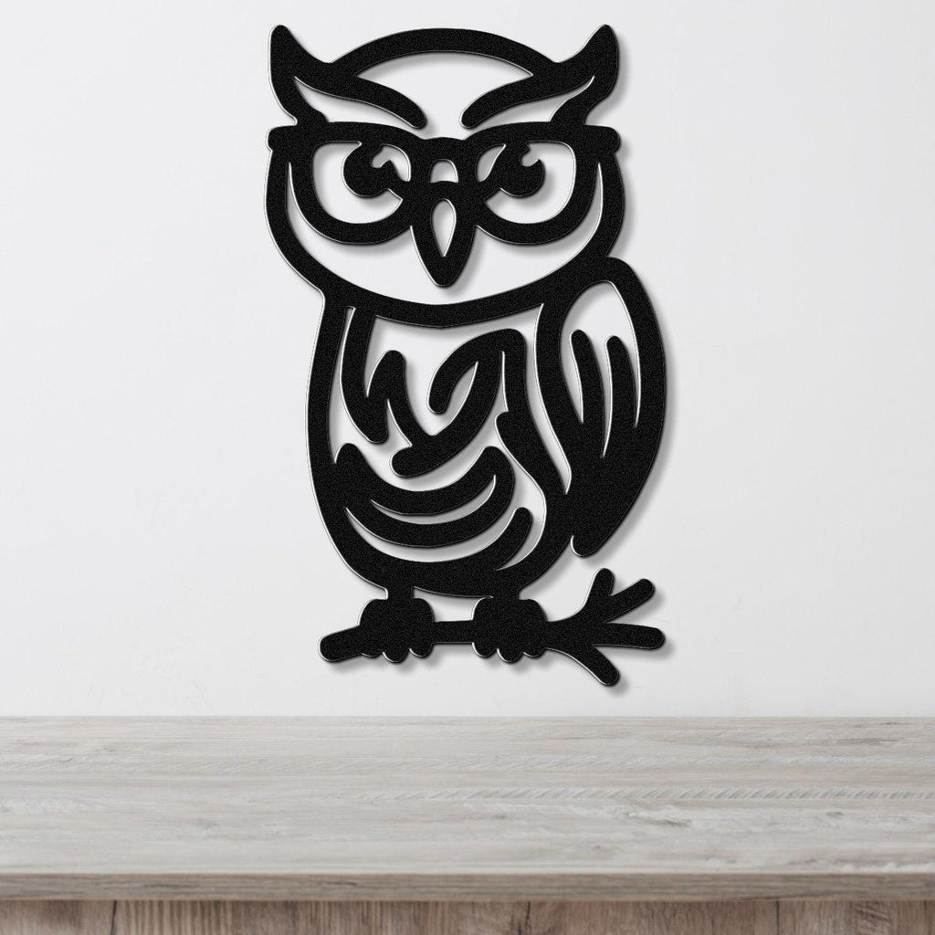 Black metal owl wall art with a bespectacled silhouette and intricate internal cutouts, mounted on a white wall above a wooden surface.