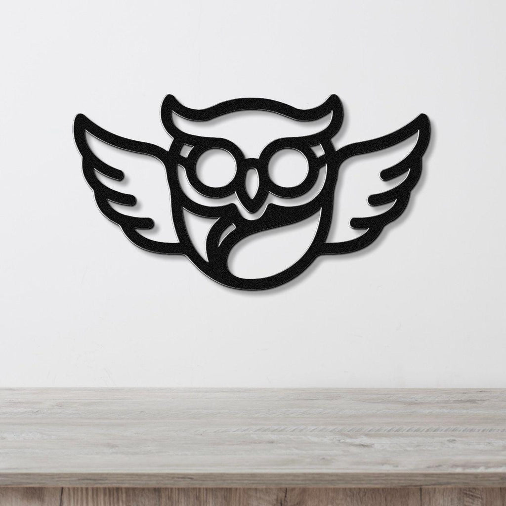 Black metal wall art featuring a bespectacled owl silhouette with wings, mounted on a white wall above a wooden surface.