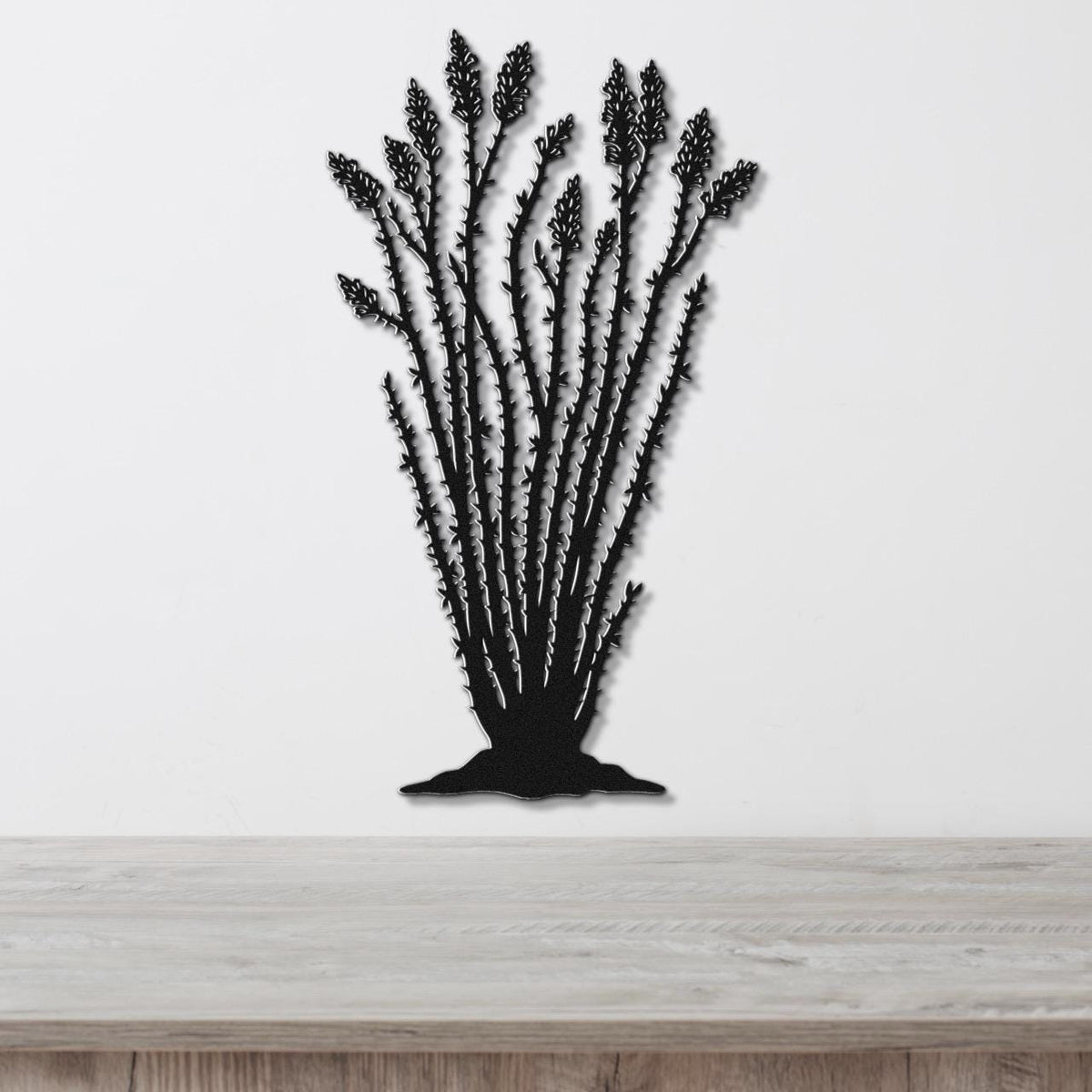 Black metal wall art silhouette of an ocotillo plant with spiny stems and clustered flower cutouts, mounted on a white wall above a light wood surface.