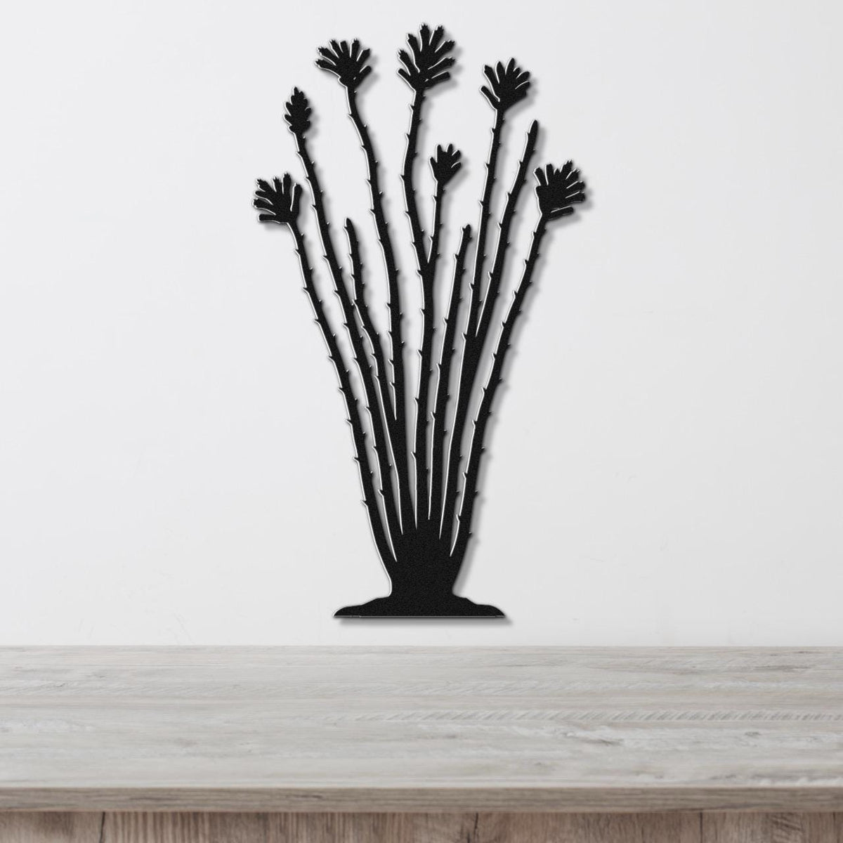 Black metal ocotillo wall sculpture with spiny stems and fan-shaped leaf cutouts, mounted on a white wall above a wooden surface.