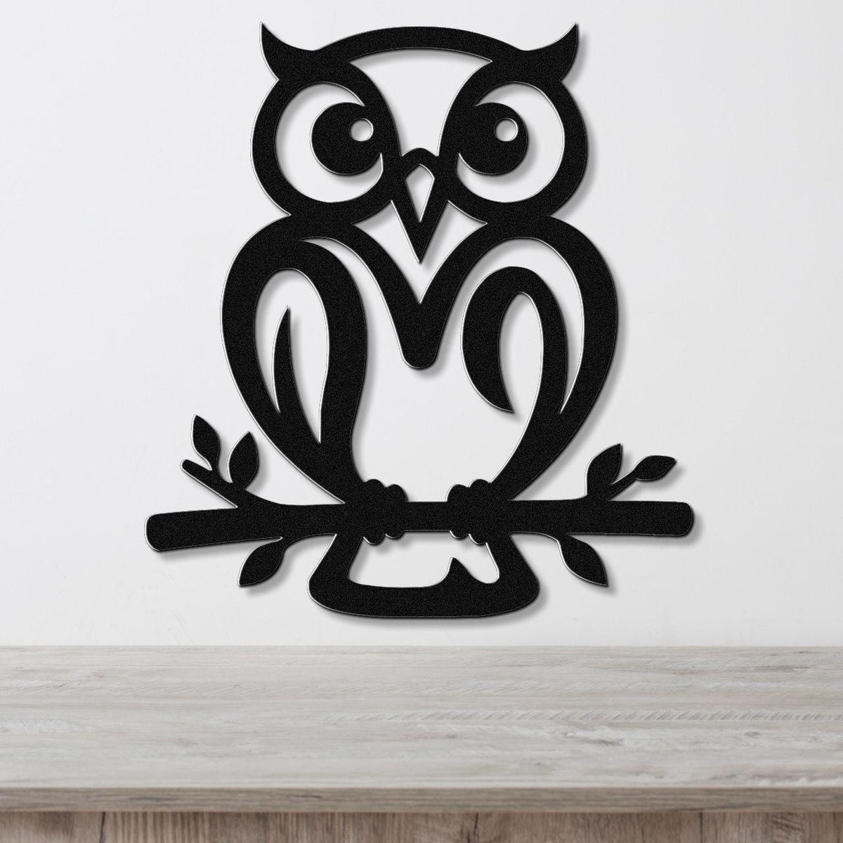 Black metal owl wall art with cutout eyes and branch details, mounted on a white wall above a wooden surface.