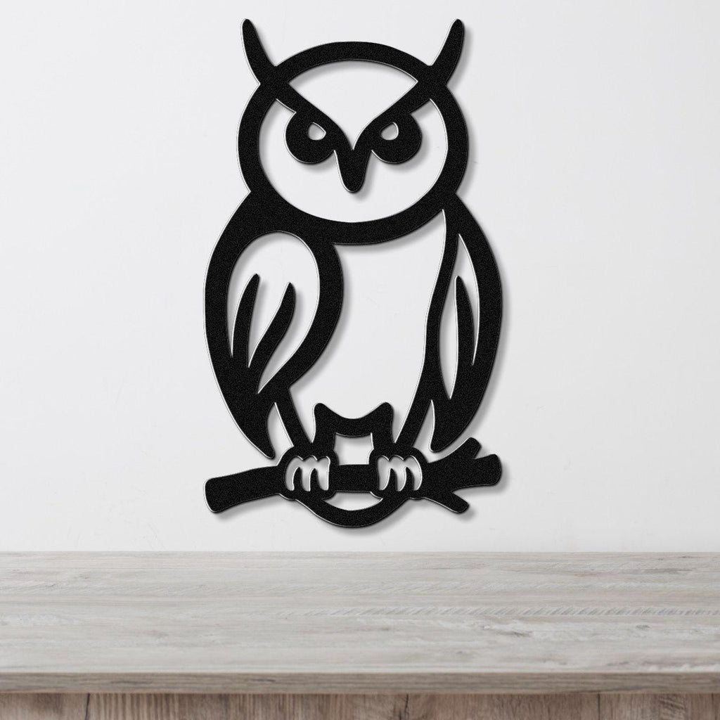 Black metal owl wall art with cutout details of eyes, ears, and talons gripping a branch, mounted on a white wall above a wooden surface.