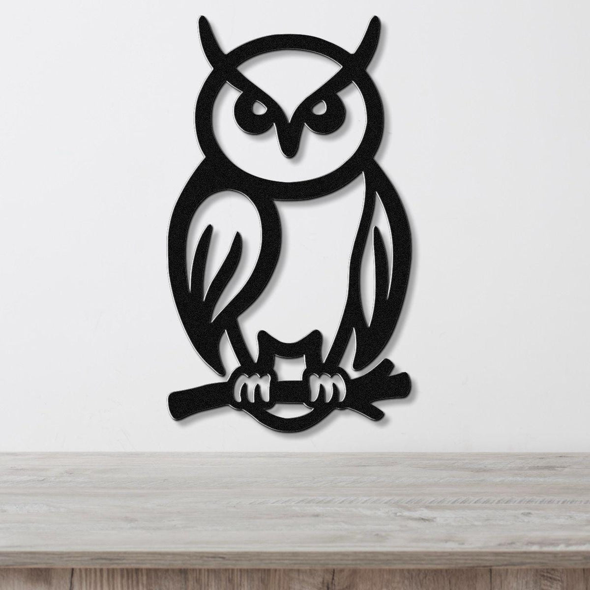 Black metal owl wall art with cutout details of eyes, ears, and talons gripping a branch, mounted on a white wall above a wooden surface.