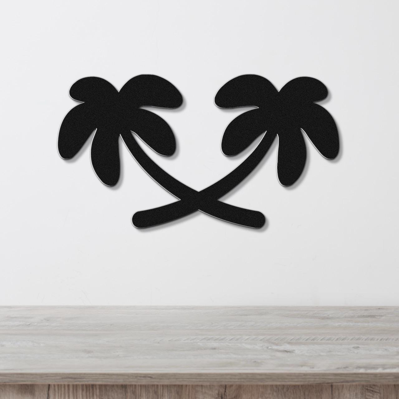 Black metal wall art featuring two palm tree silhouettes with crossed trunks, mounted on a white wall above a light wood surface.