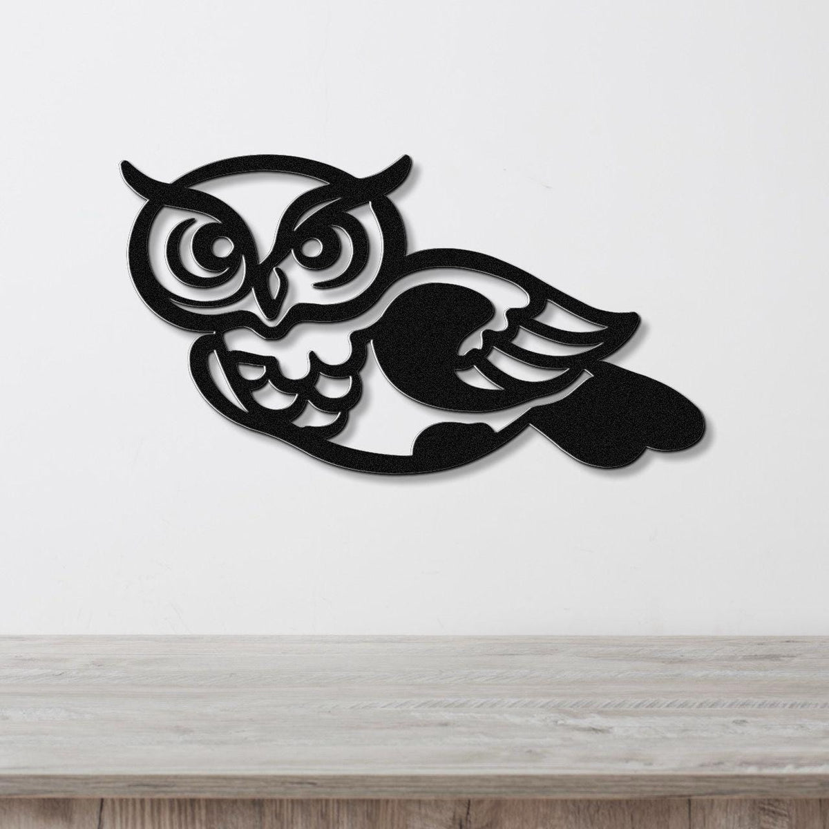 Black metal owl wall art with intricate cutout details of eyes, wings, and feathers, mounted on a white wall above a light wood surface.