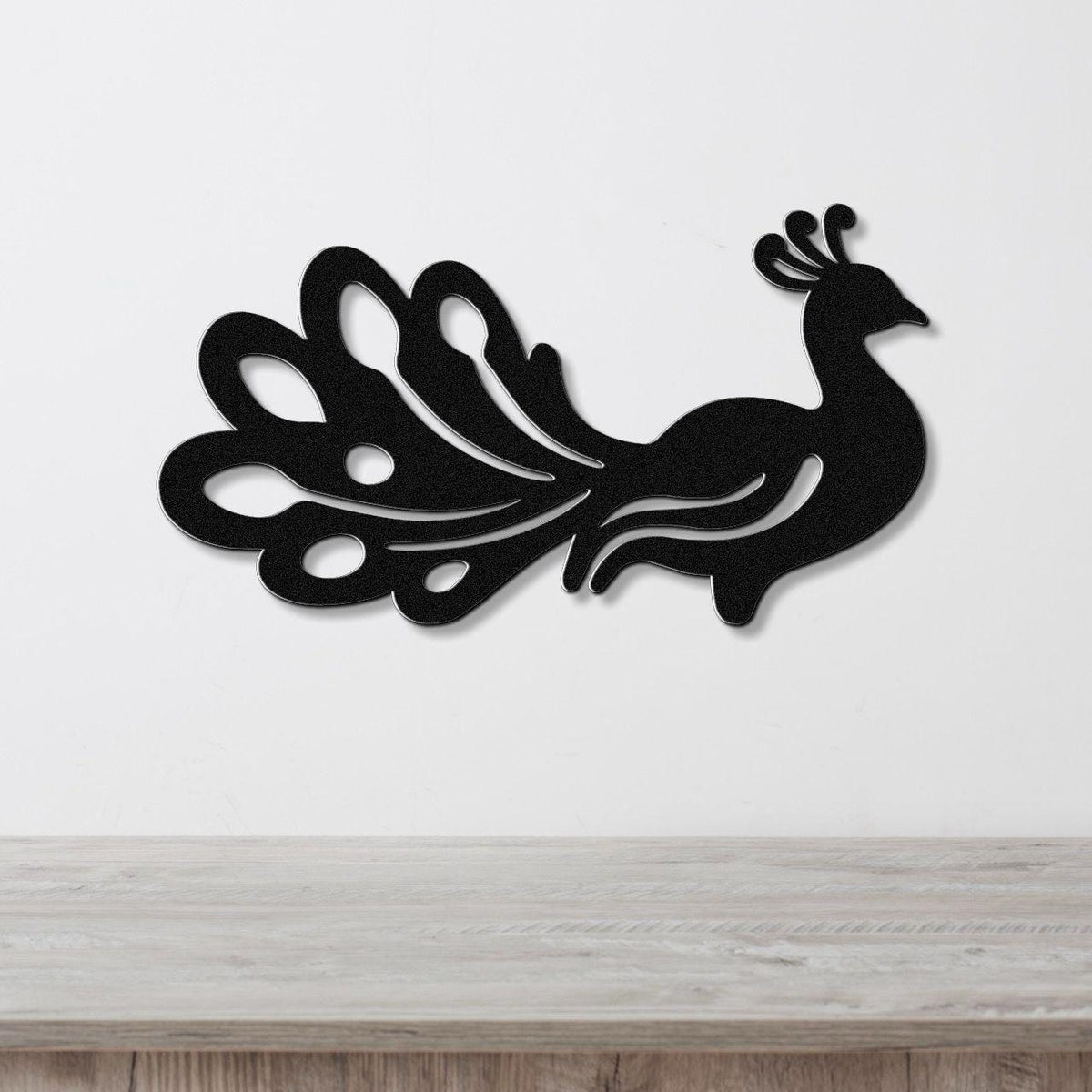 Black metal peacock wall art with a silhouette design and cutout tail feathers, mounted on a white wall above a wooden surface.