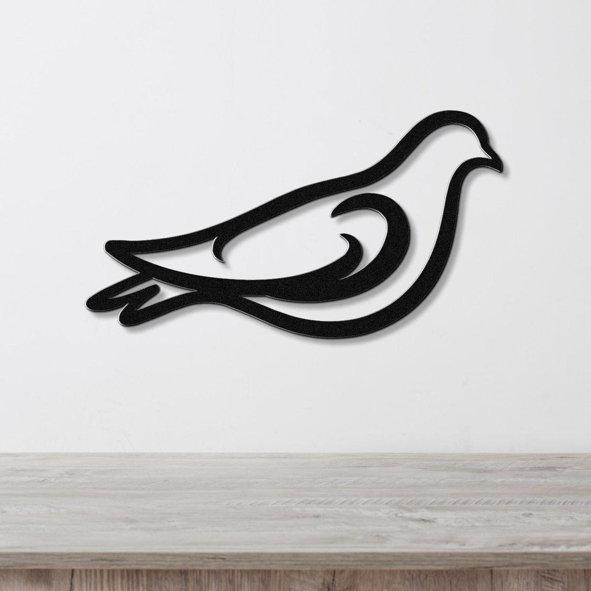 Black metal pigeon wall art with a bold silhouette and internal cutout details, mounted on a white wall above a light wood surface.