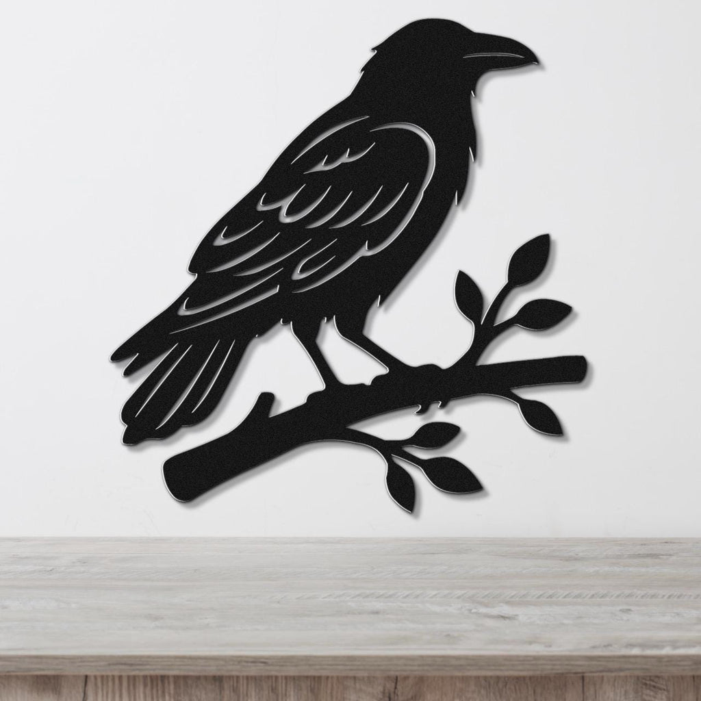 Black metal raven wall art silhouette perched on a branch with leaf cutouts, mounted on a white wall above a light wood surface.