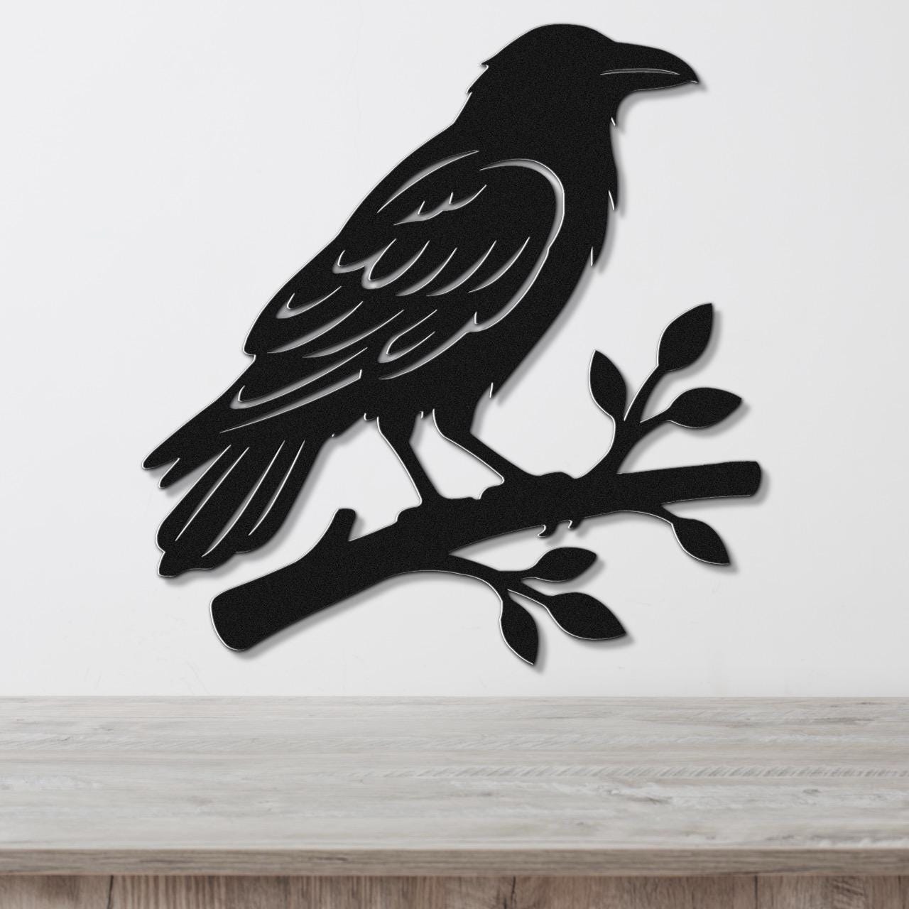 Black metal raven wall art silhouette perched on a branch with leaf cutouts, mounted on a white wall above a light wood surface.