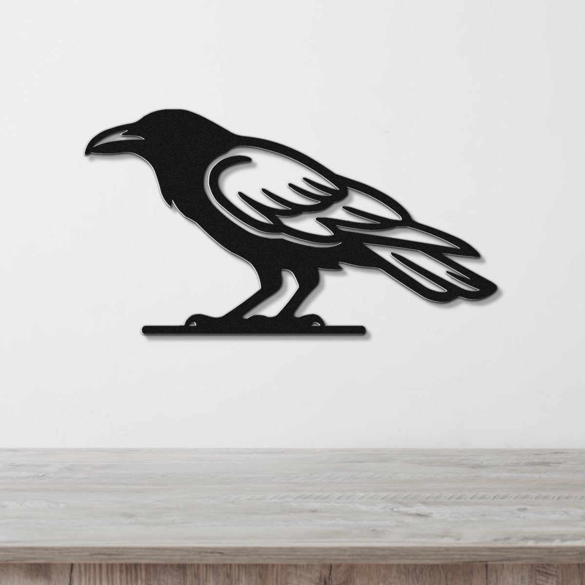 Black metal raven wall art with cutout wing and tail details, mounted on a white wall above a light wood surface.
