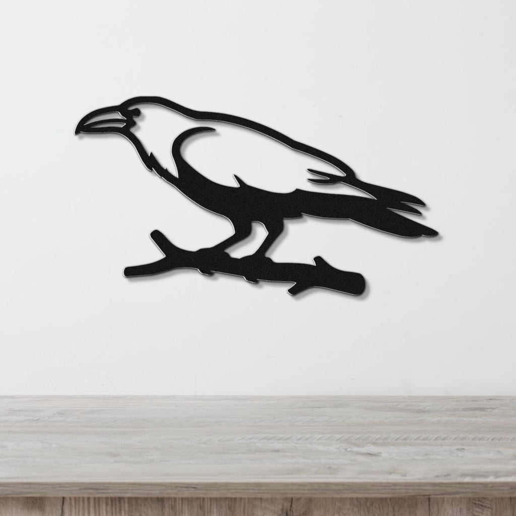 Black metal raven wall art silhouette perched on a branch, mounted on a white wall above a light wood surface.