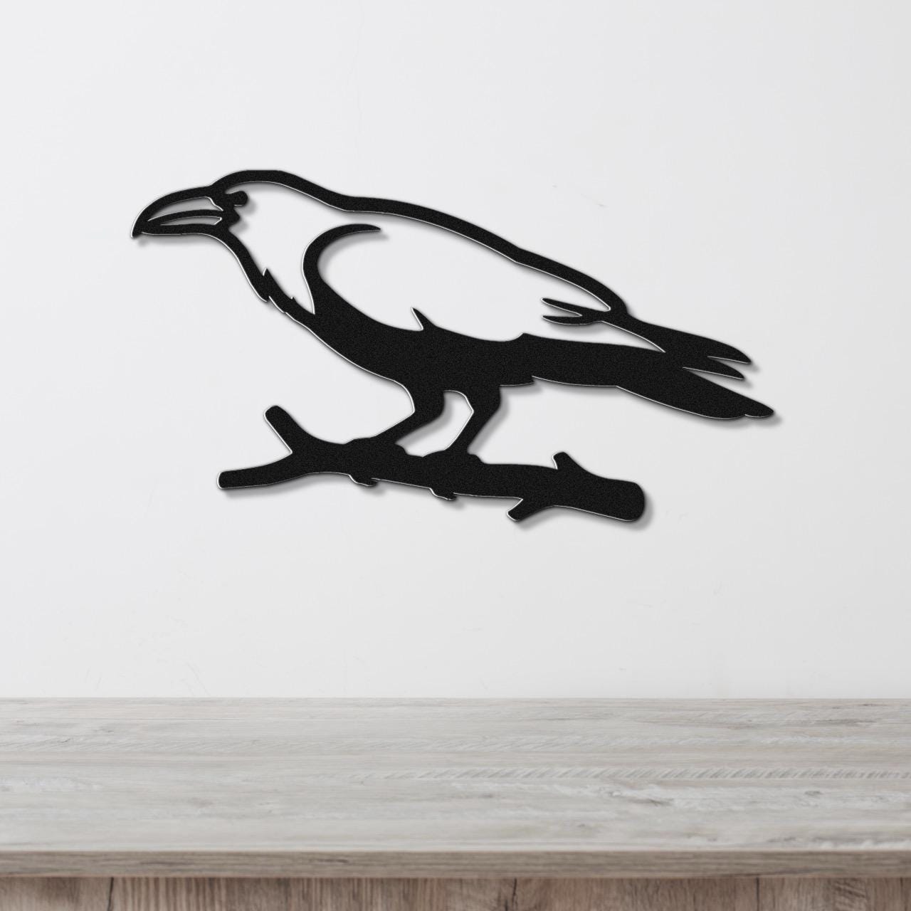 Black metal raven wall art silhouette perched on a branch, mounted on a white wall above a light wood surface.