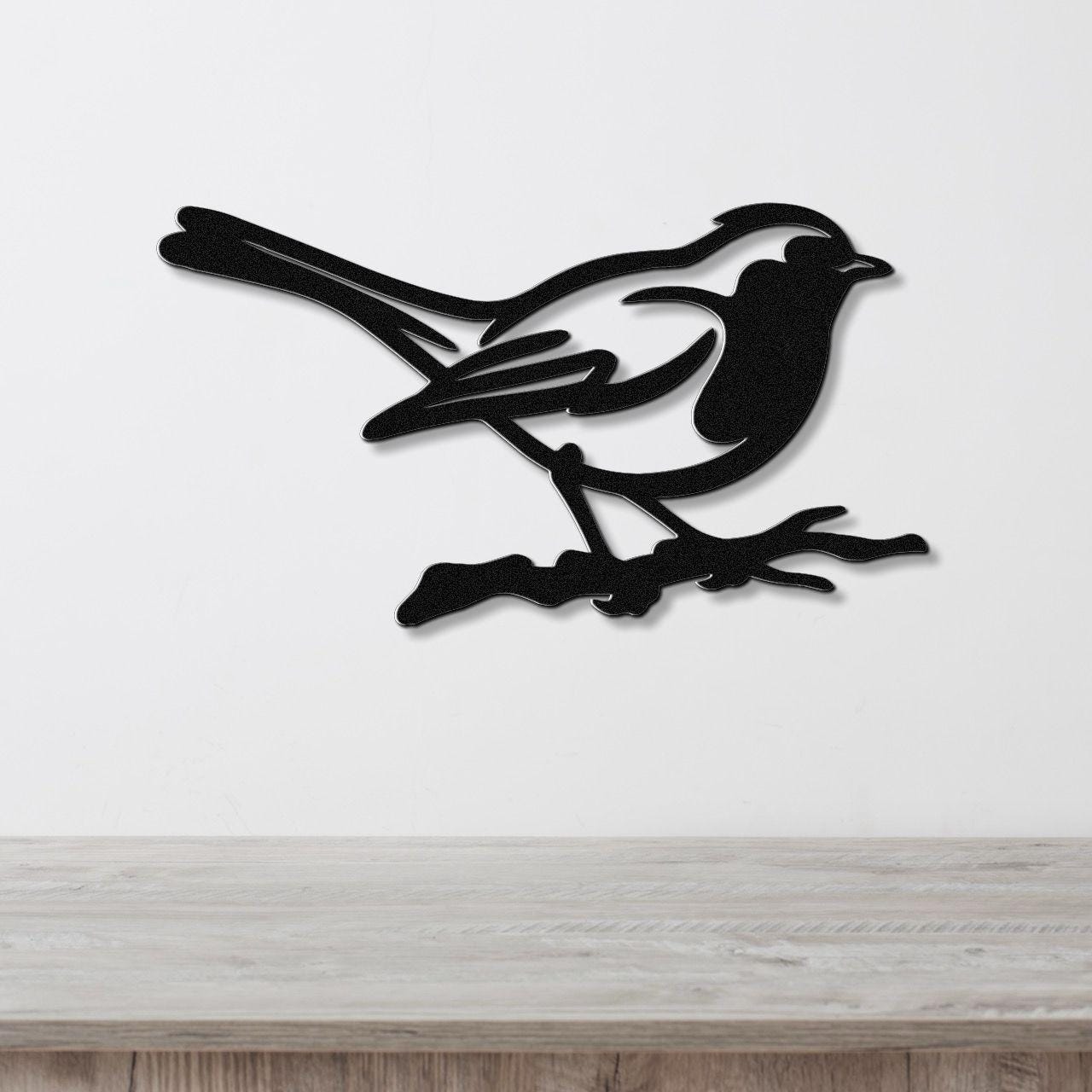 Black metal bird wall art in a minimalist silhouette style, perched on a branch, mounted on a white wall above a light wood surface.