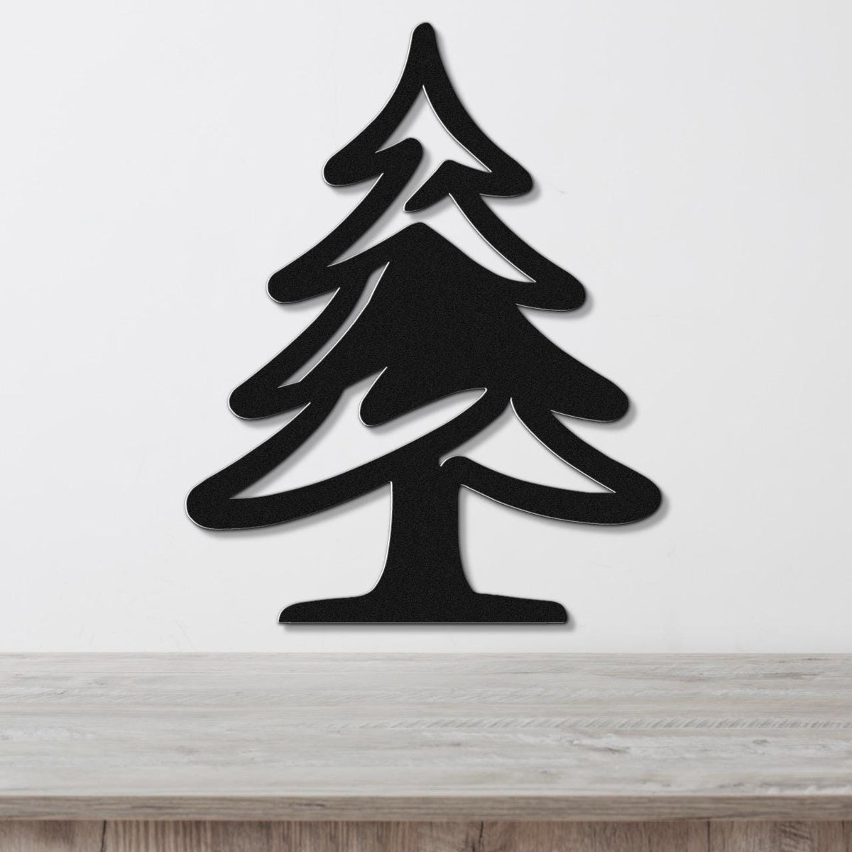 Black metal pine tree wall art with layered branch cutouts, mounted on a white wall above a light wood surface.