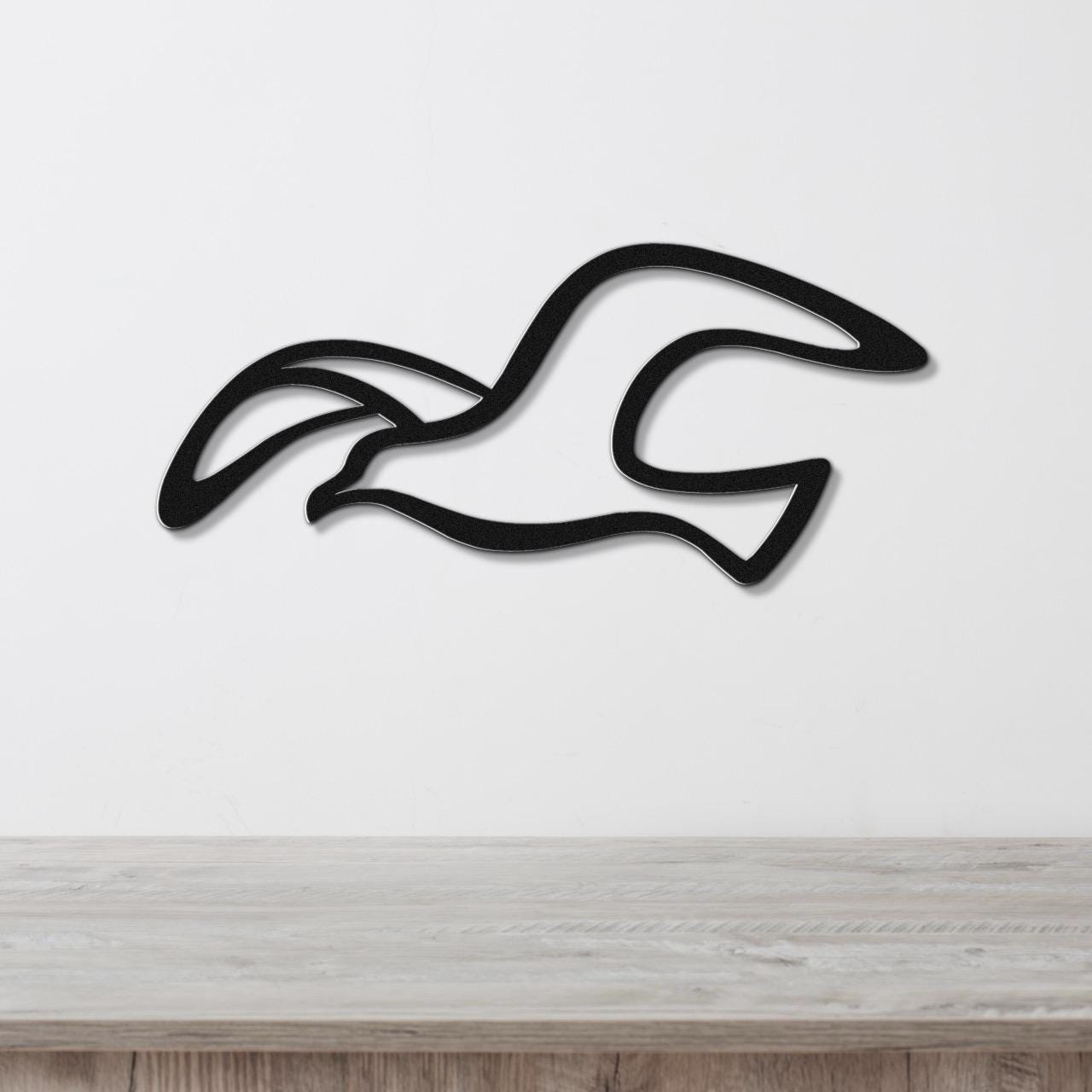 Black metal seagull wall art with flowing wing cutouts, mounted on a white wall above a light wood surface.