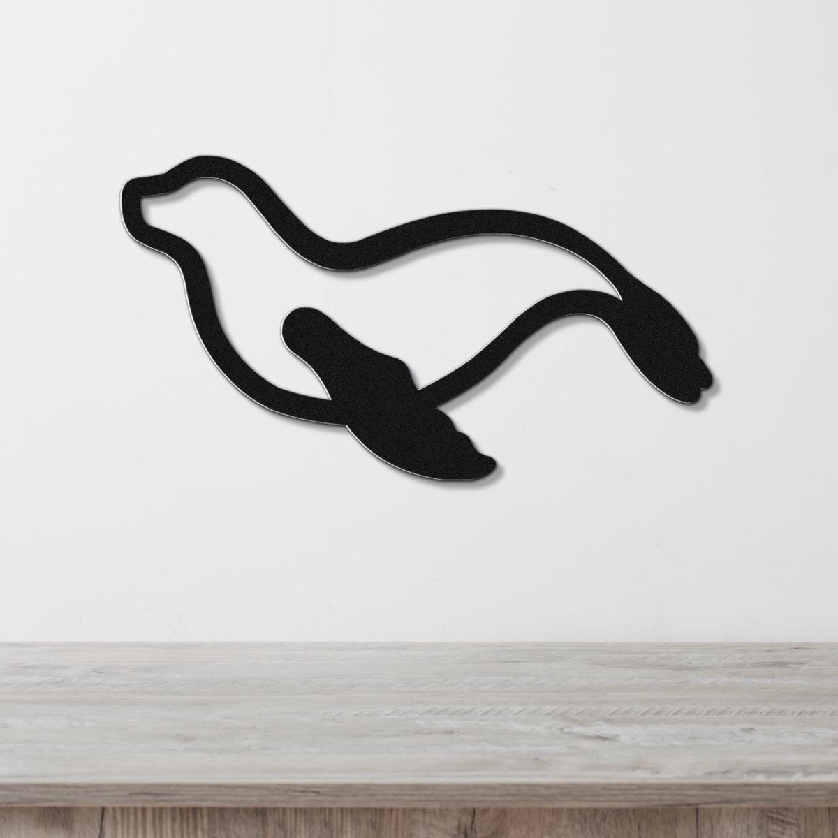 Black metal sea lion wall art with a flowing silhouette design, mounted on a white wall above a light wood surface.