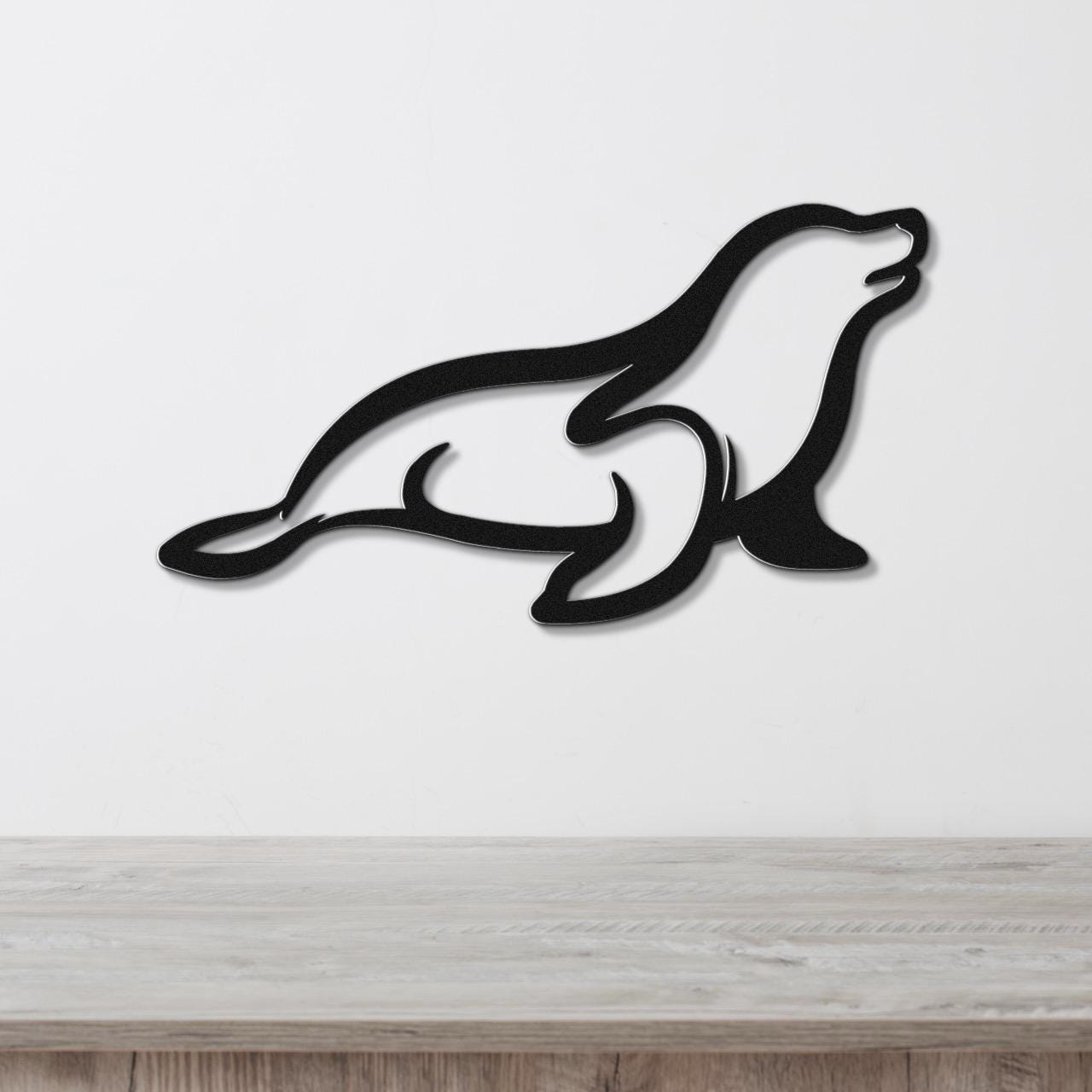Black metal sea lion wall art with a sleek silhouette design, mounted on a white wall above a light wood surface.