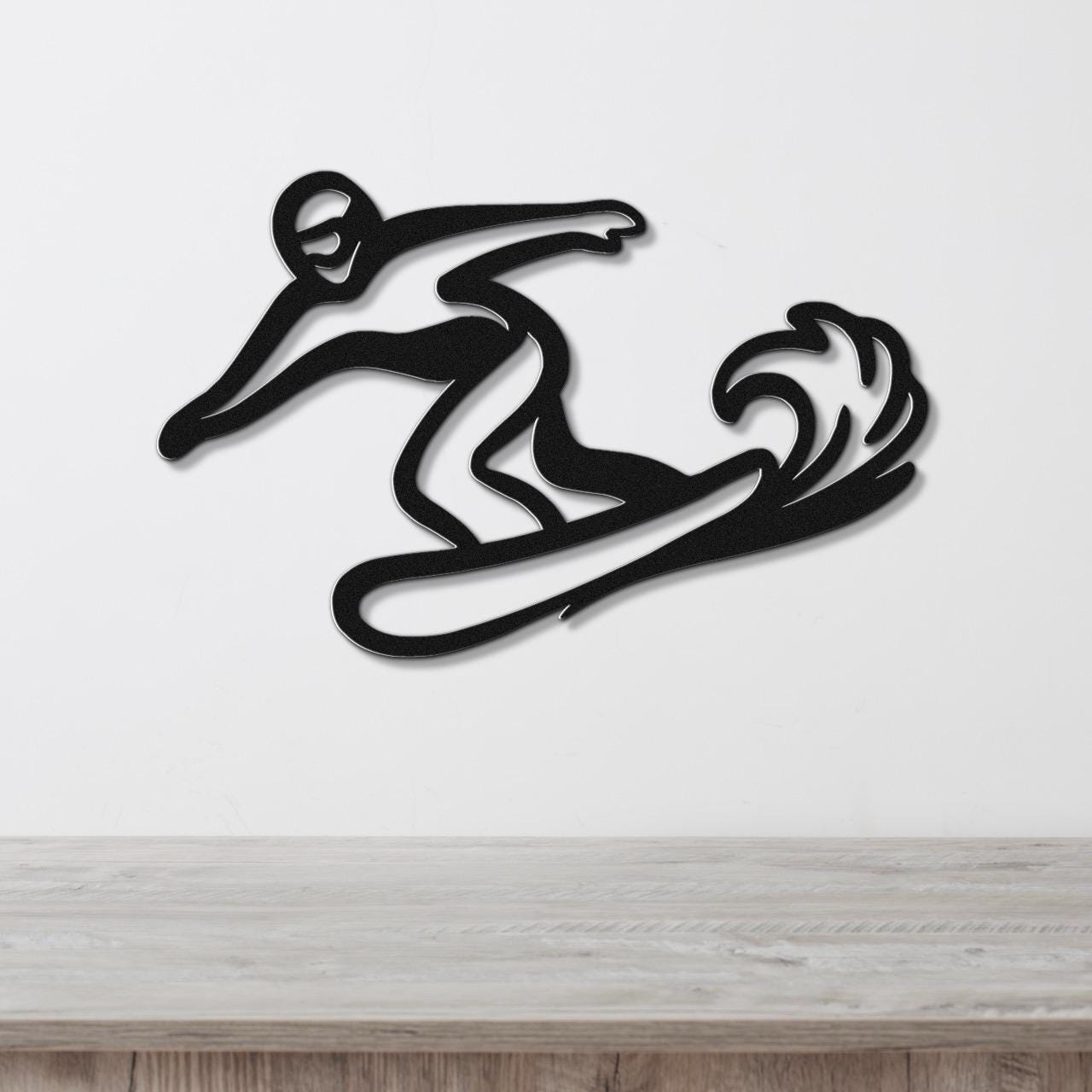 Black metal wall art silhouette of a snowboarder in motion with wave-like cutout detail, mounted on a white wall above a wooden surface.