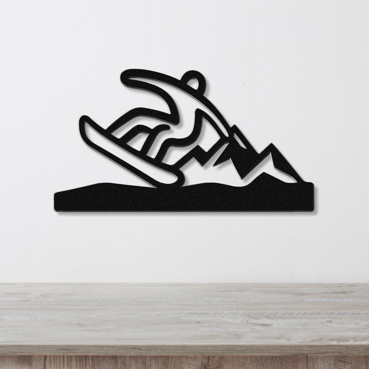 Black metal wall sculpture of a snowboarder in motion, with cutout details of mountains and snow, mounted on a white wall above a wooden surface.