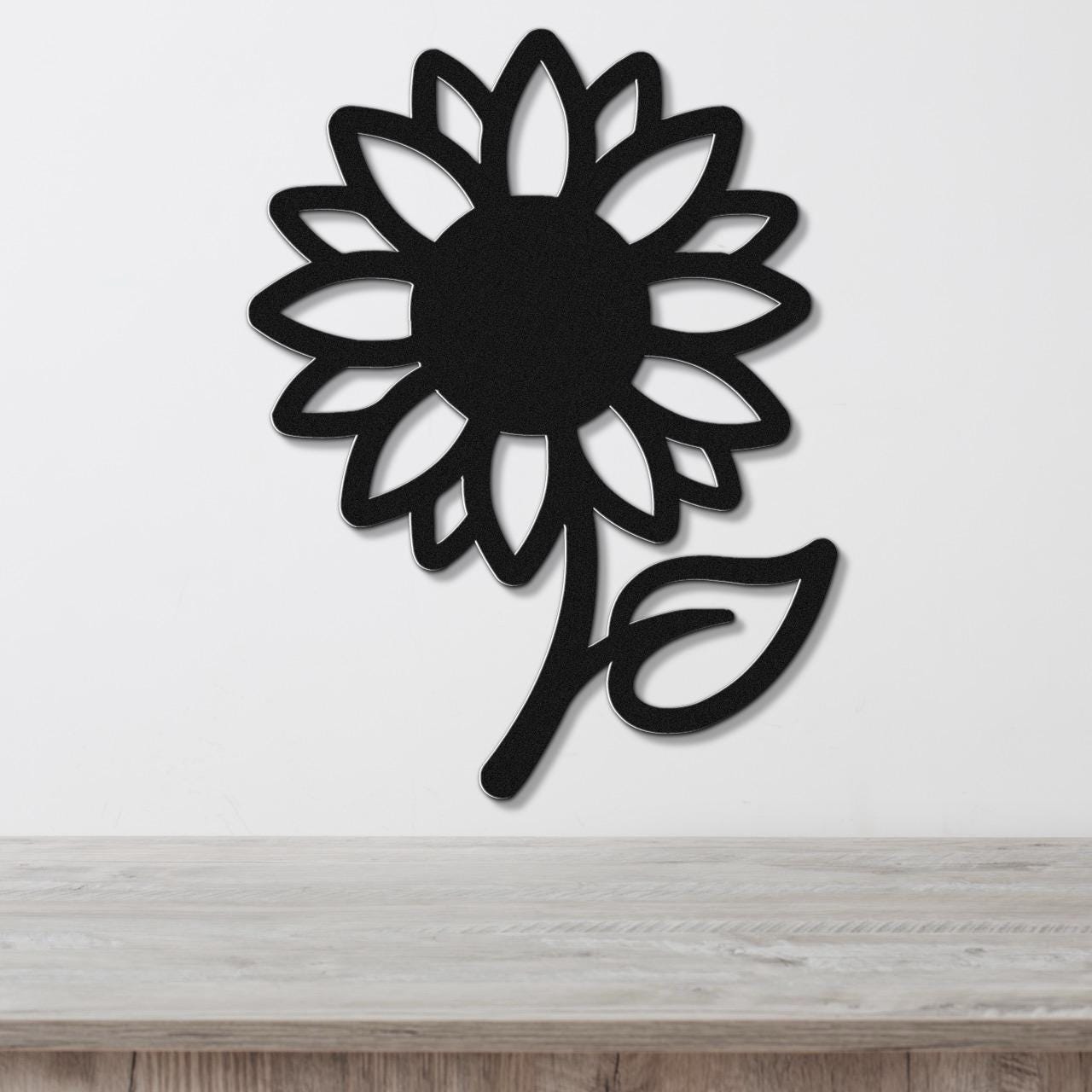 Black metal sunflower wall art with a bold silhouette and cutout petals, mounted on a white wall above a light wood countertop.